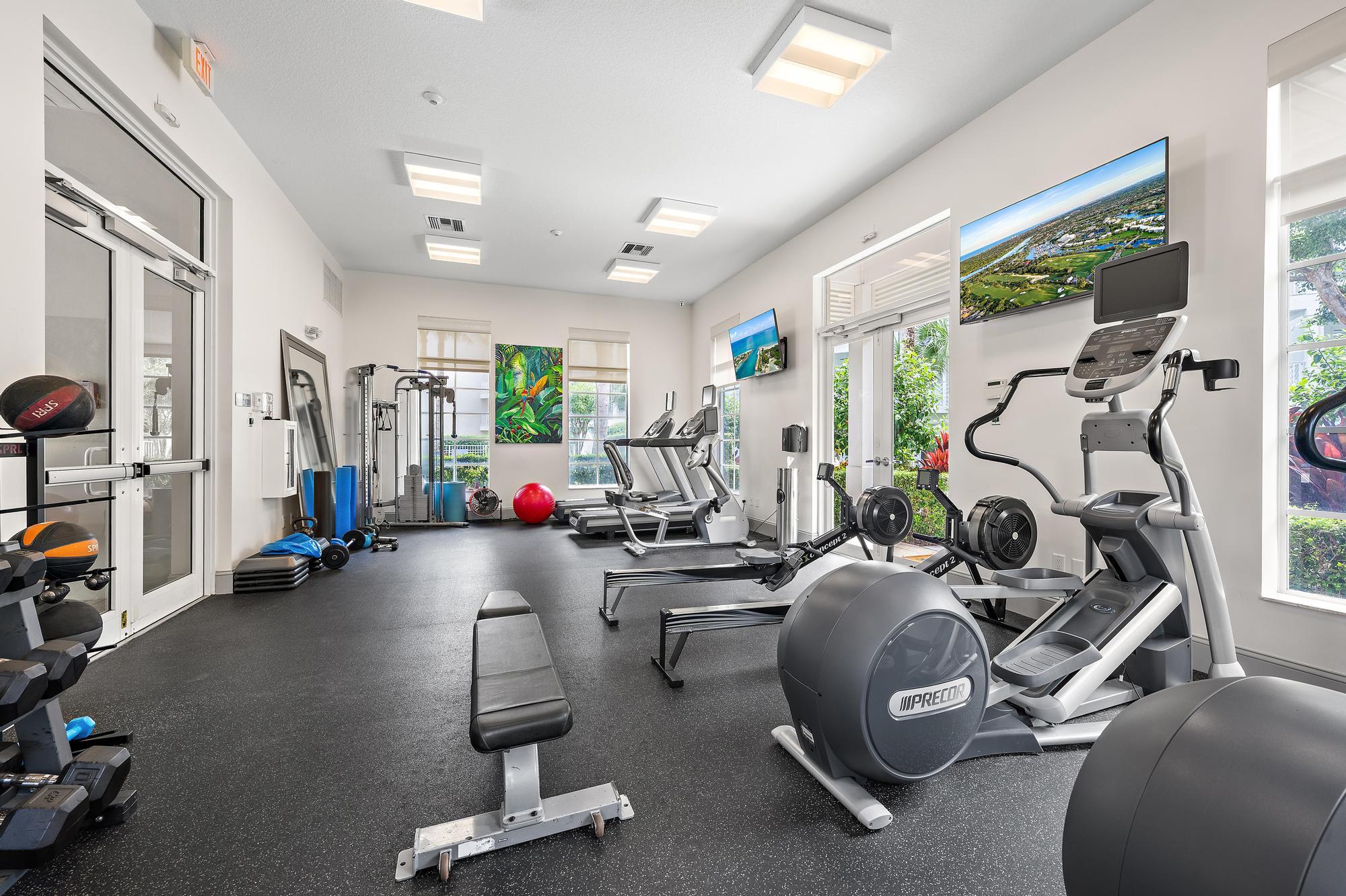 Fitness room with rows of cardio machines, weight equipment, and large windows overlooking greenery.