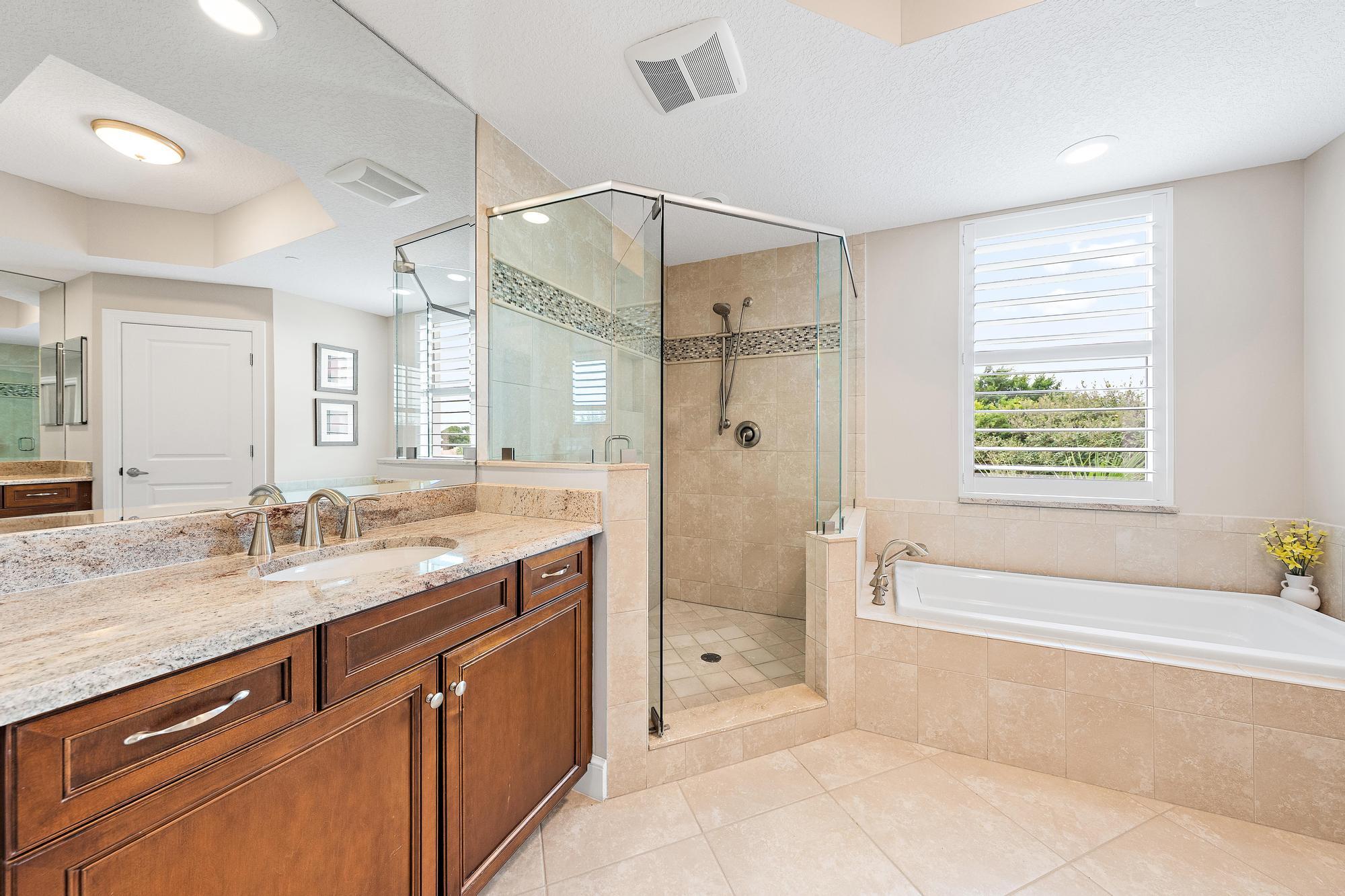 Bathroom with a granite double vanity, glass shower, and built-in tub beneath plantation shutters.