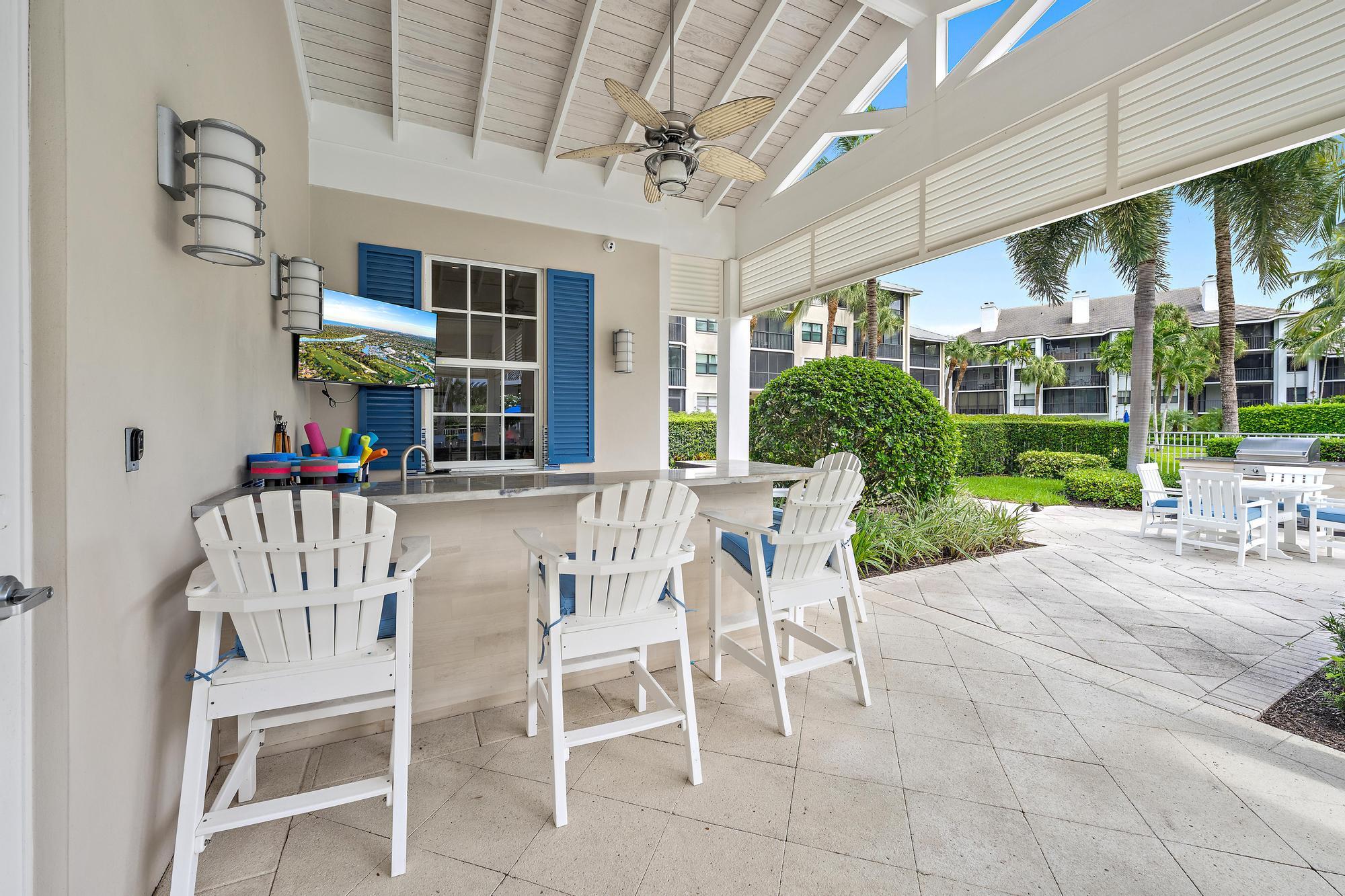 Covered patio with a bar counter, white Adirondack chairs, and a ceiling fan overlooking palm trees.