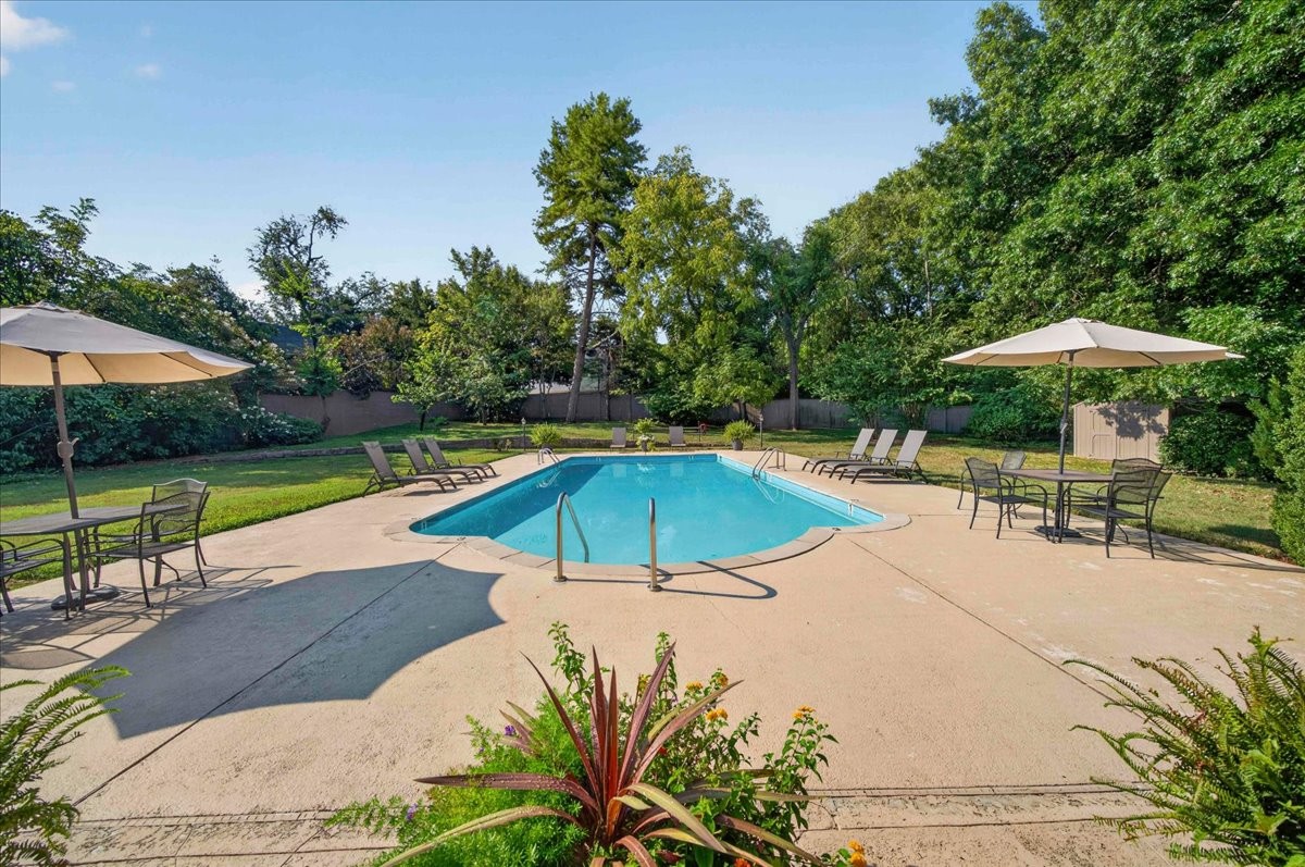 Outdoor pool with an umbrella-shaded patio, lounge chairs, and mature trees.