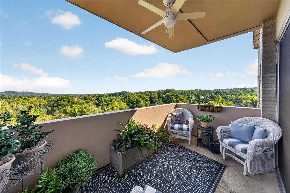 Furnished balcony with wicker seating and potted plants overlooking wooded hills.