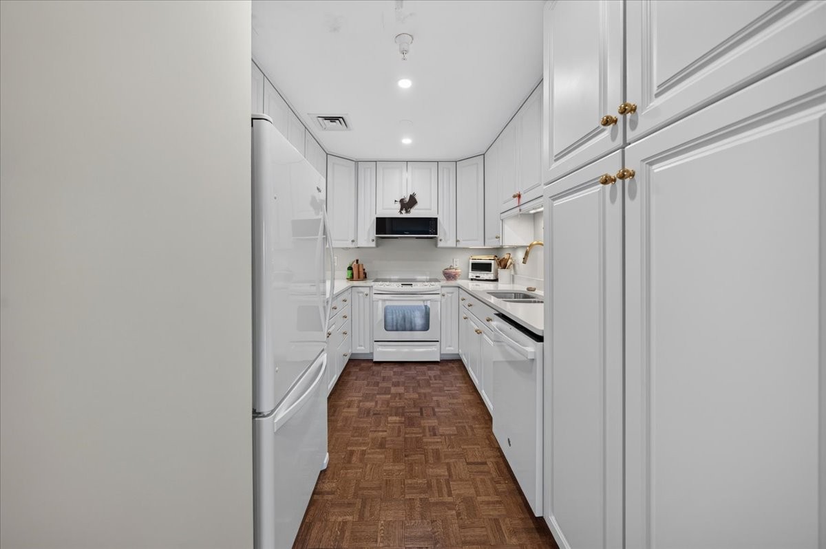 Galley kitchen with white cabinetry and appliances, featuring a brown parquet floor.