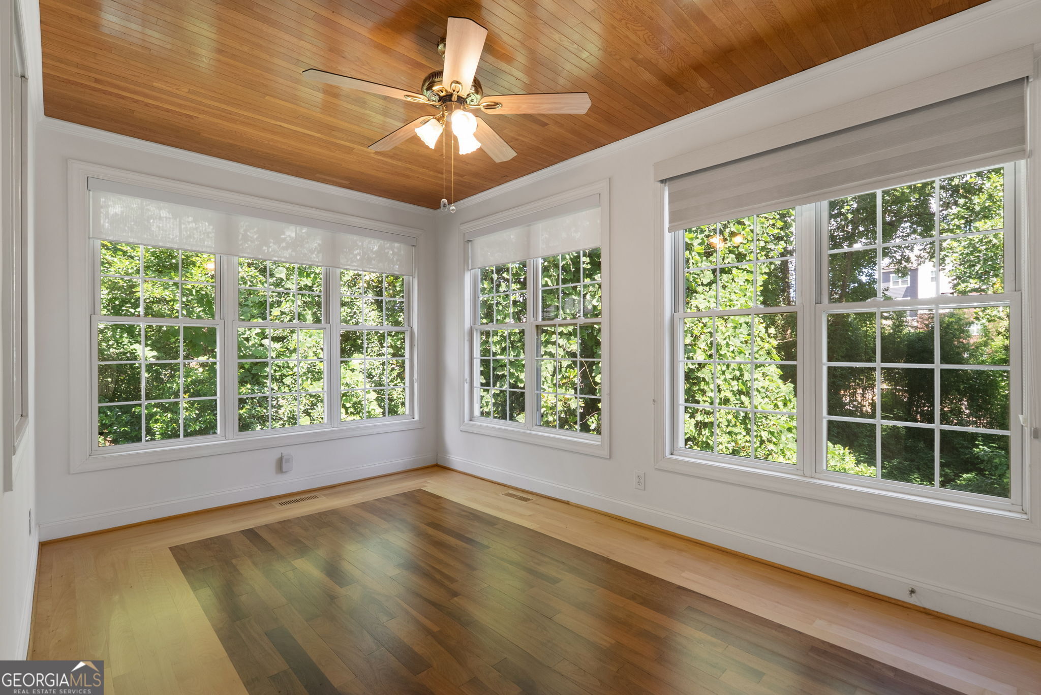 Empty sunroom with a wood-plank ceiling, hardwood floors, and large windows overlooking greenery.