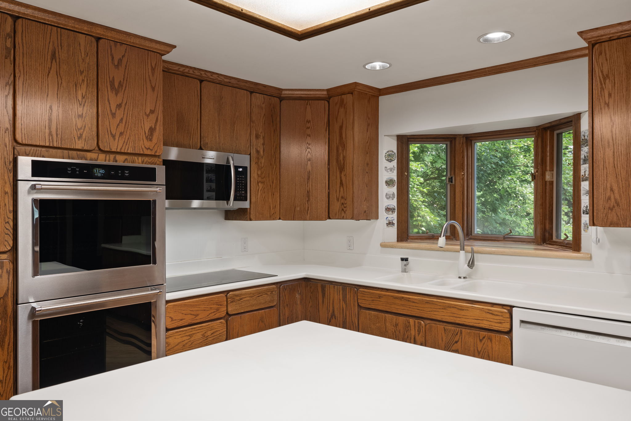 Kitchen with oak cabinets, stainless-steel double ovens, and a sink beneath a leafy bay window.