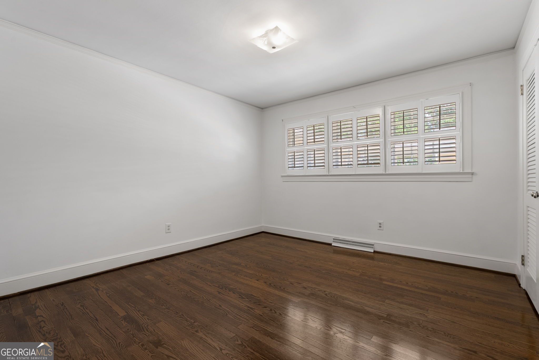 Empty bedroom with dark hardwood floors and plantation shutters over the windows.