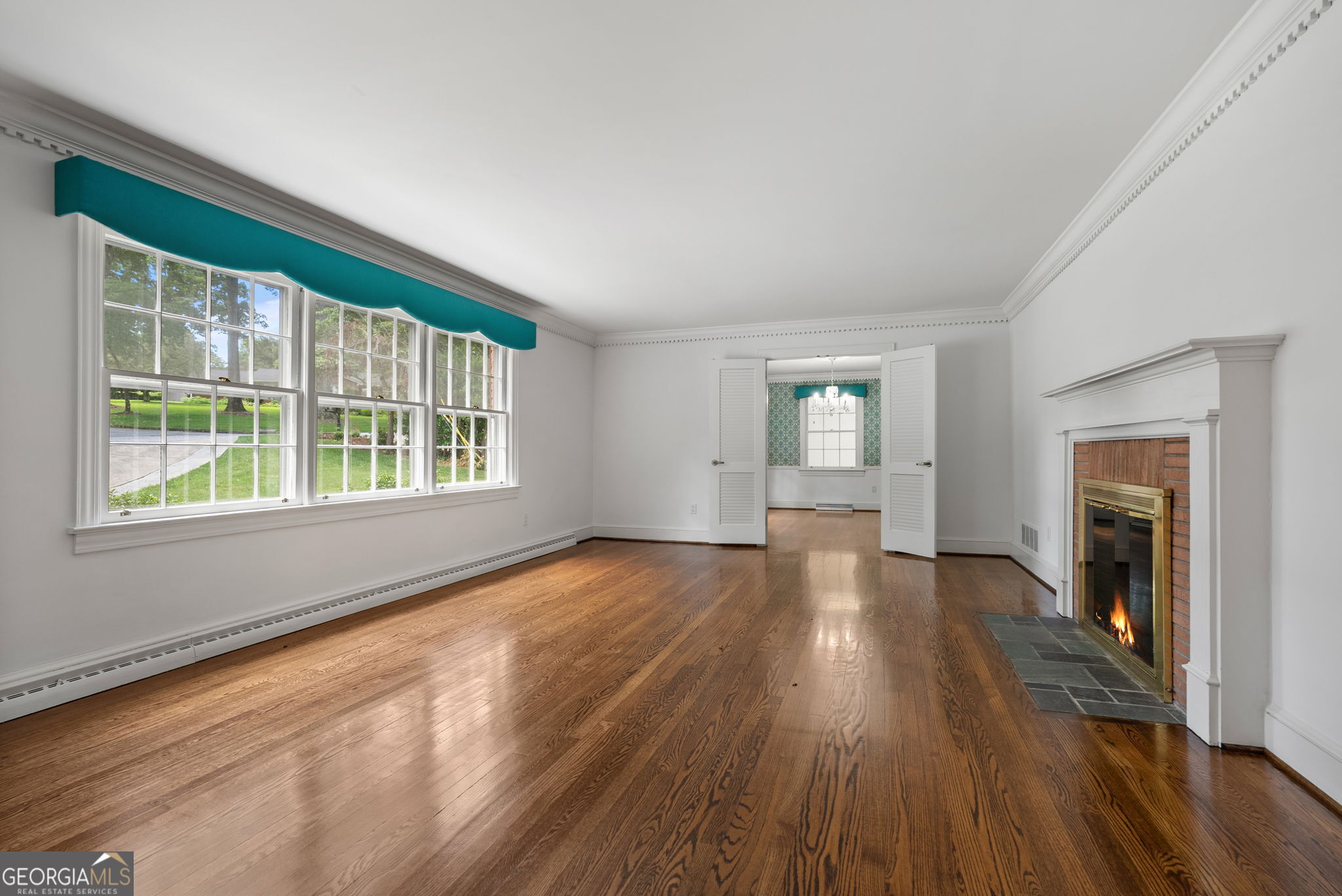 Vacant living room with hardwood floors, a brick fireplace, and large windows topped with teal valances.