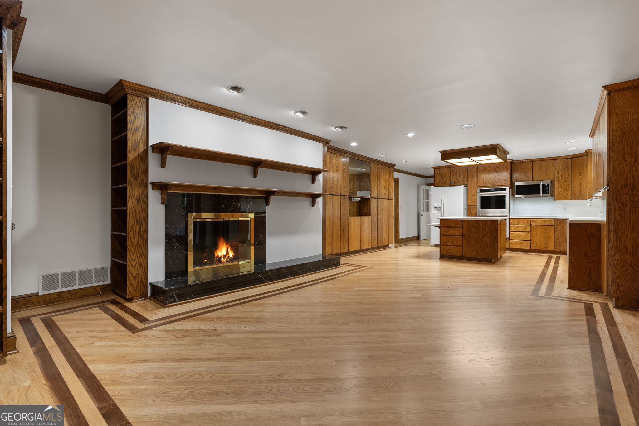 Open kitchen and living area with warm wood cabinetry, patterned wood flooring, and a lit fireplace.