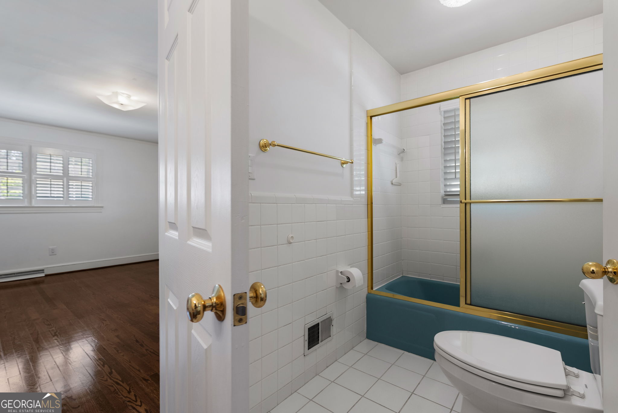 White-tiled bathroom with a teal bathtub, gold-framed shower doors, and a view into a room with hardwood floors.