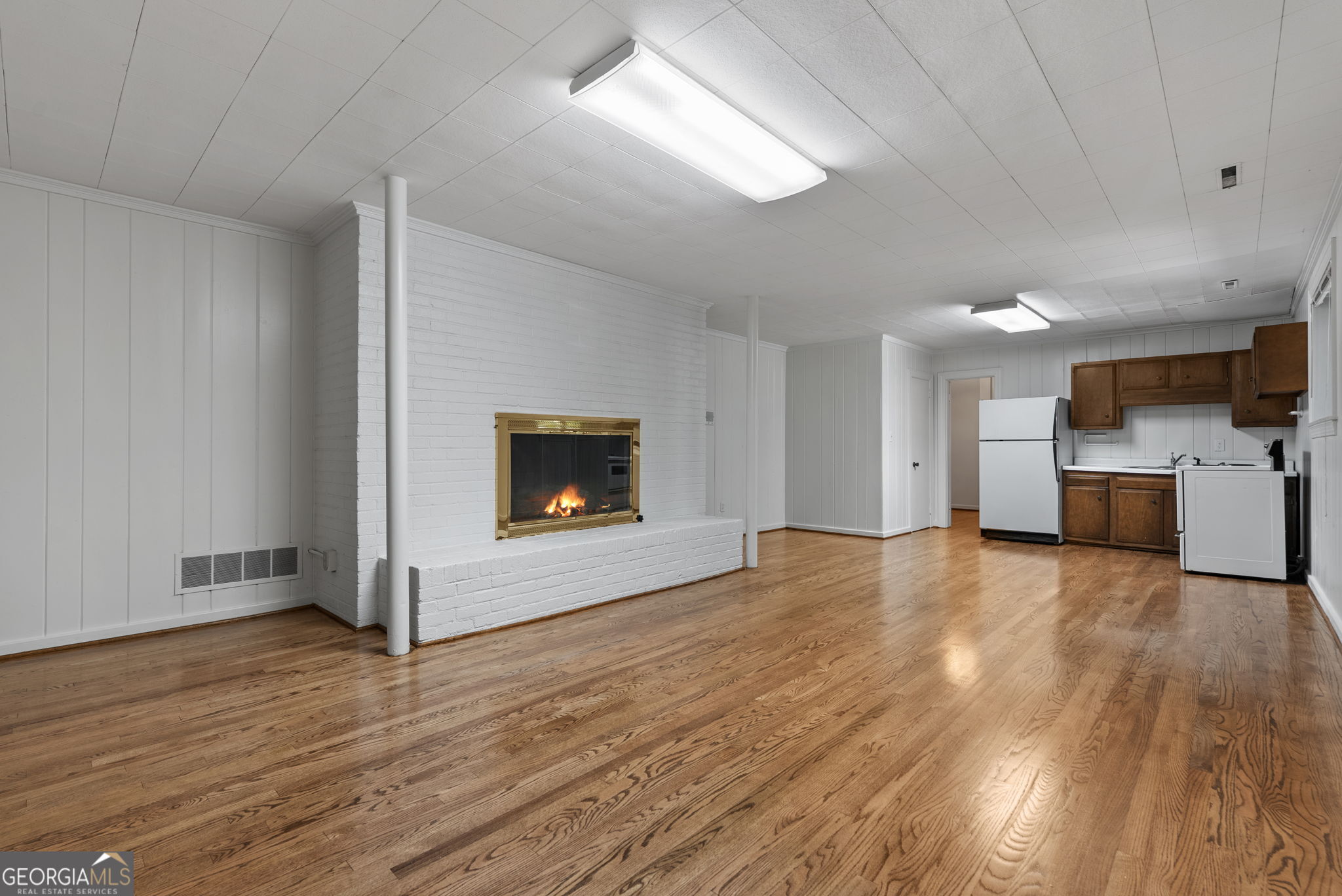 Vacant room with hardwood floors, a white brick fireplace, and a compact kitchen with wood cabinets and appliances.