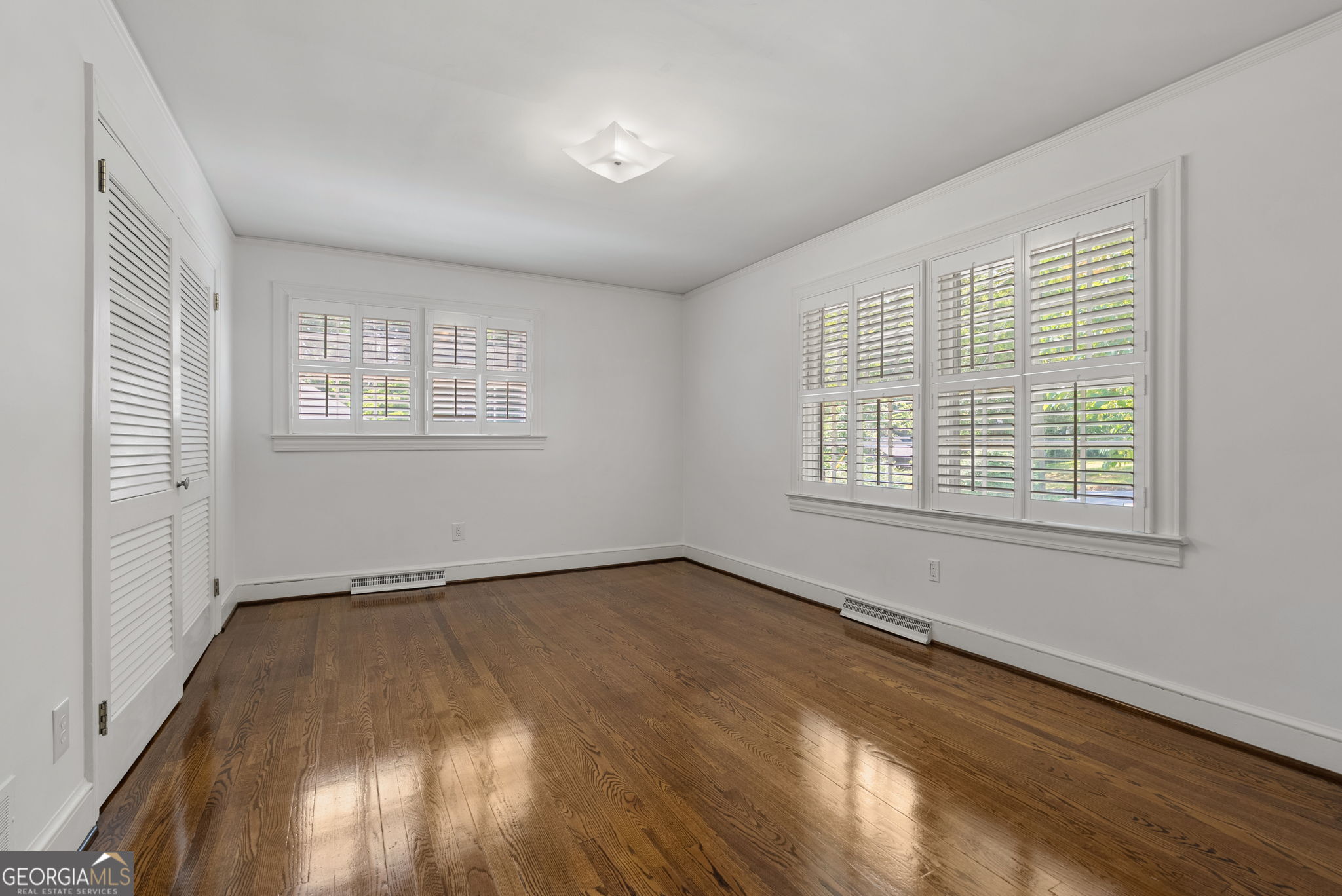 Empty bedroom with polished hardwood floors, plantation shutters, and louvered closet doors.