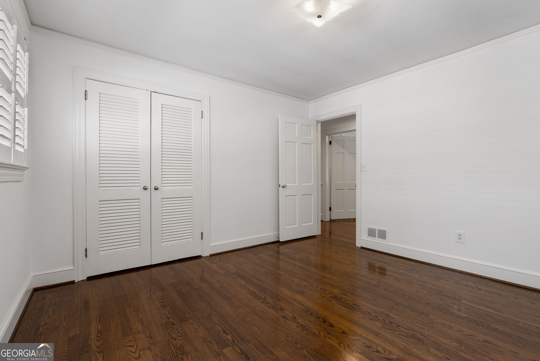 Empty bedroom with dark hardwood floors, white louvered closet doors, and an open doorway.