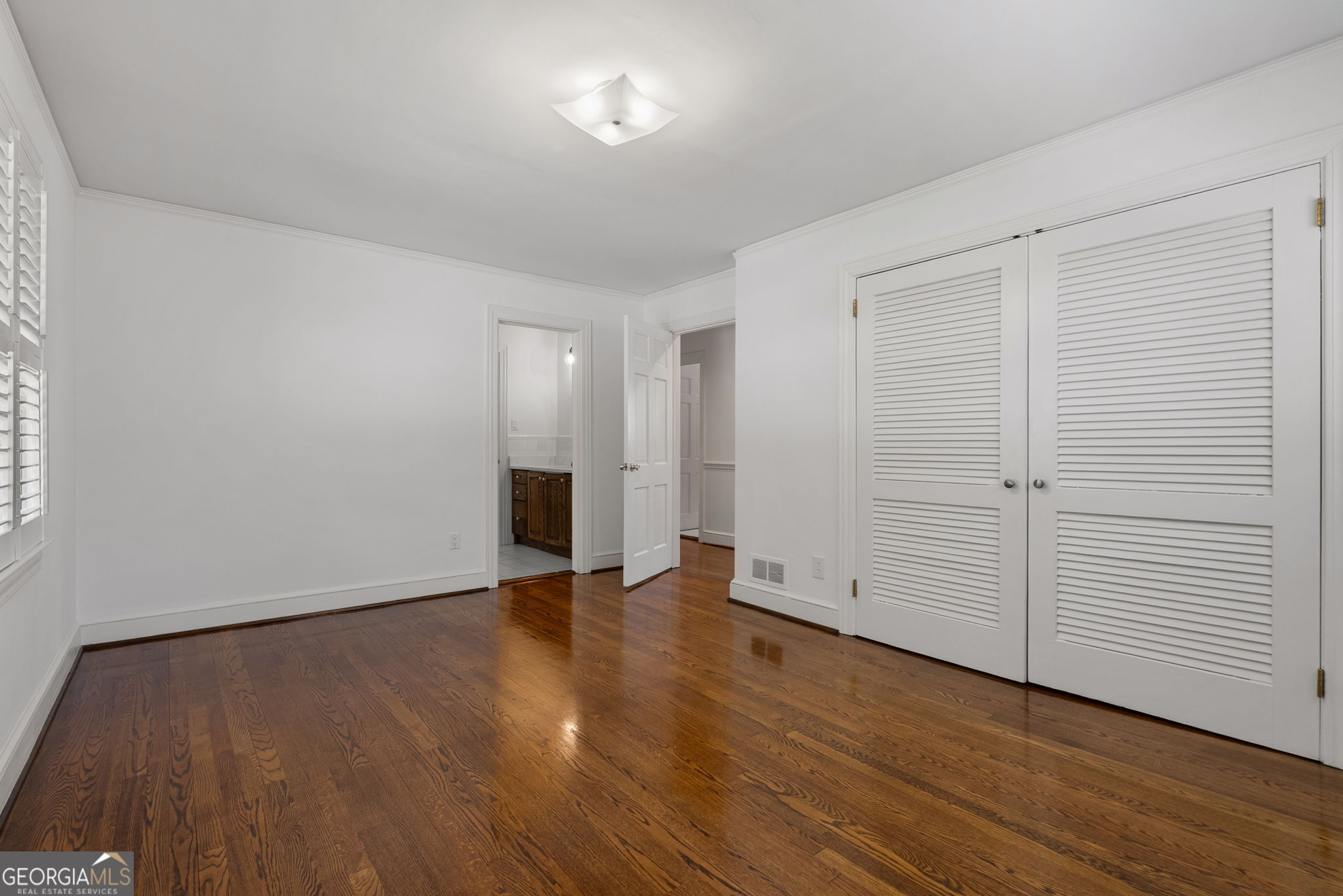 Empty bedroom with hardwood floors, plantation shutters, and louvered closet doors.