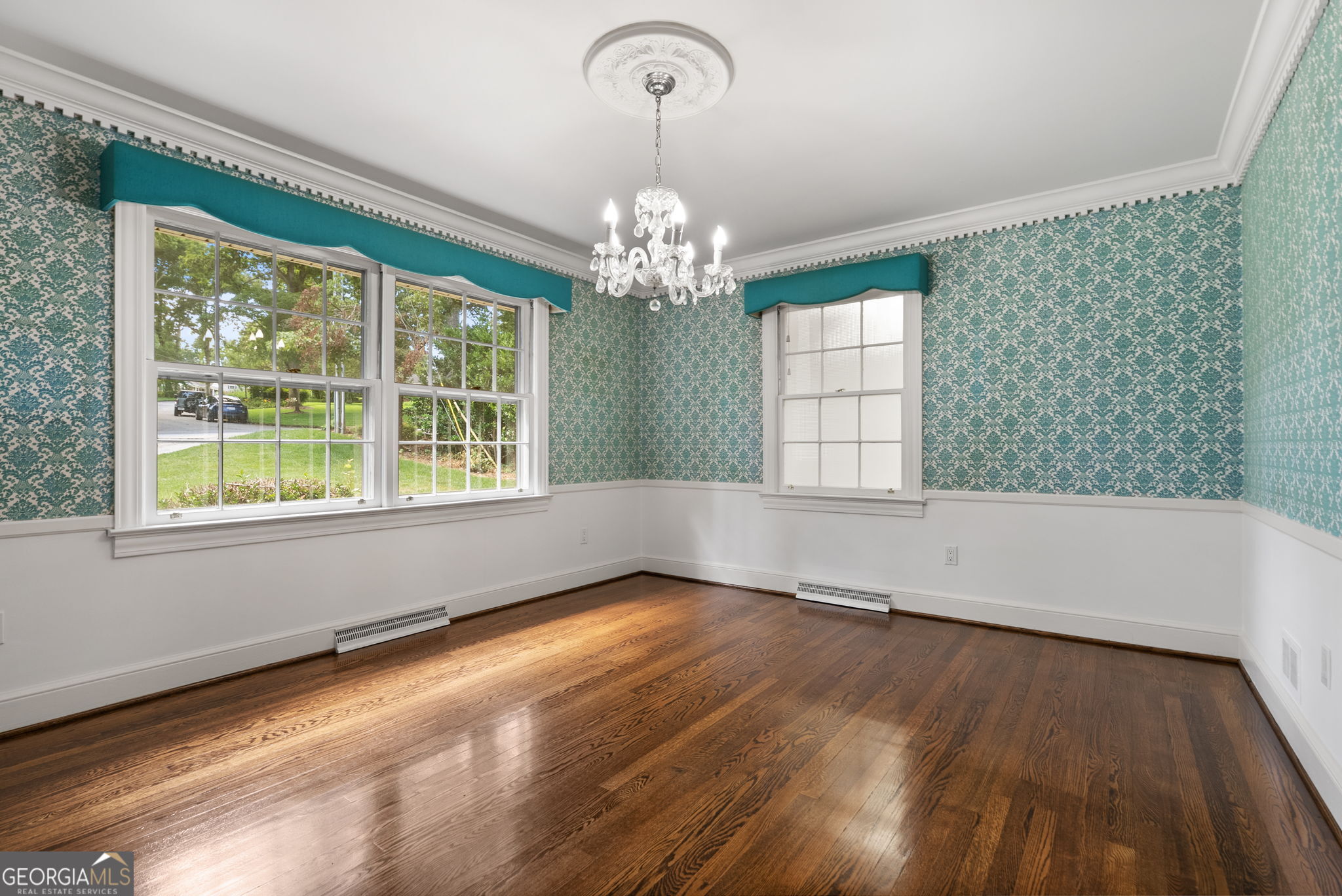 Empty room with teal patterned wallpaper, polished hardwood floors, large windows, and a crystal chandelier.