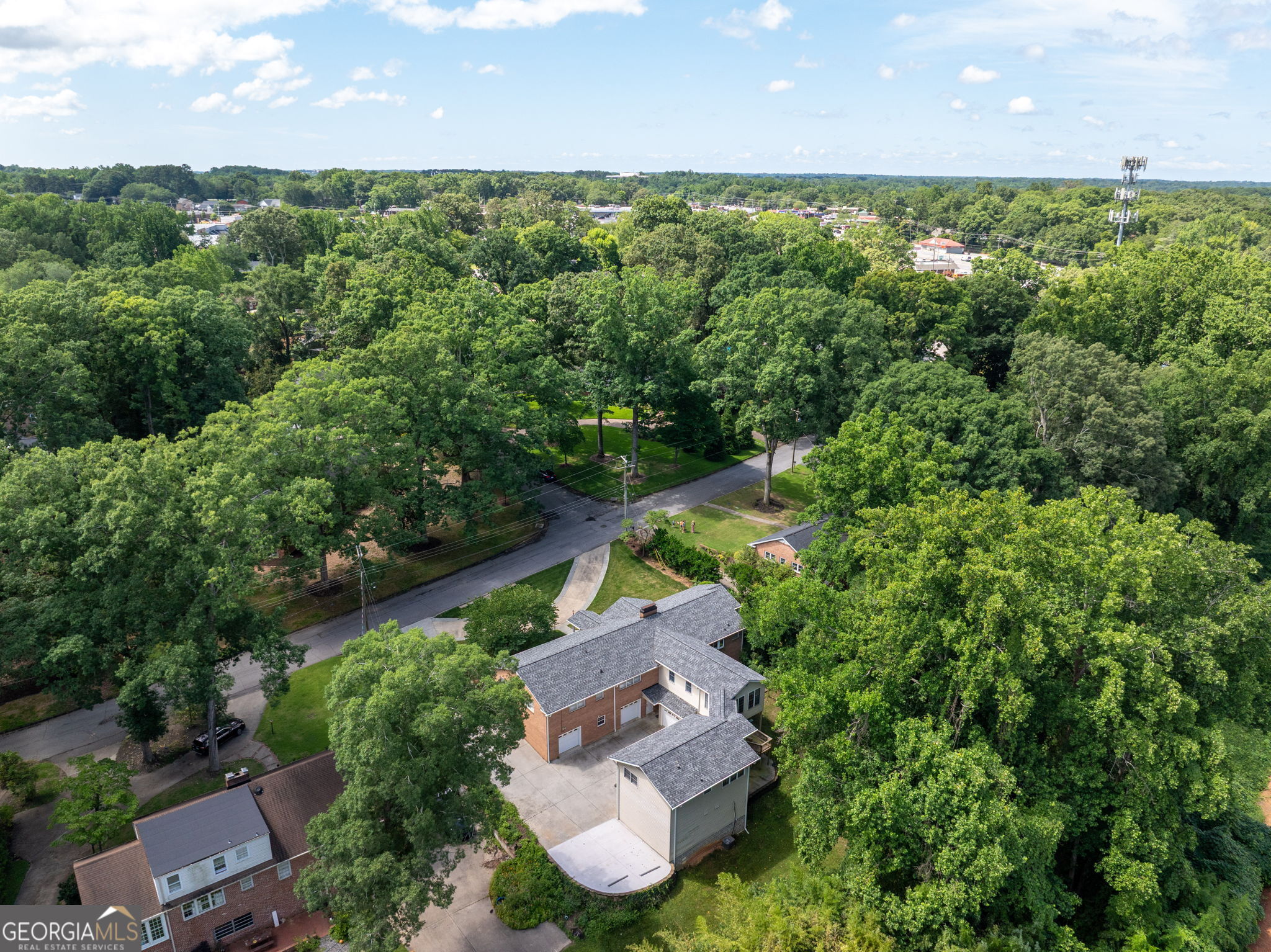 Aerial view of a brick-and-siding home amid mature trees, with a residential road alongside.