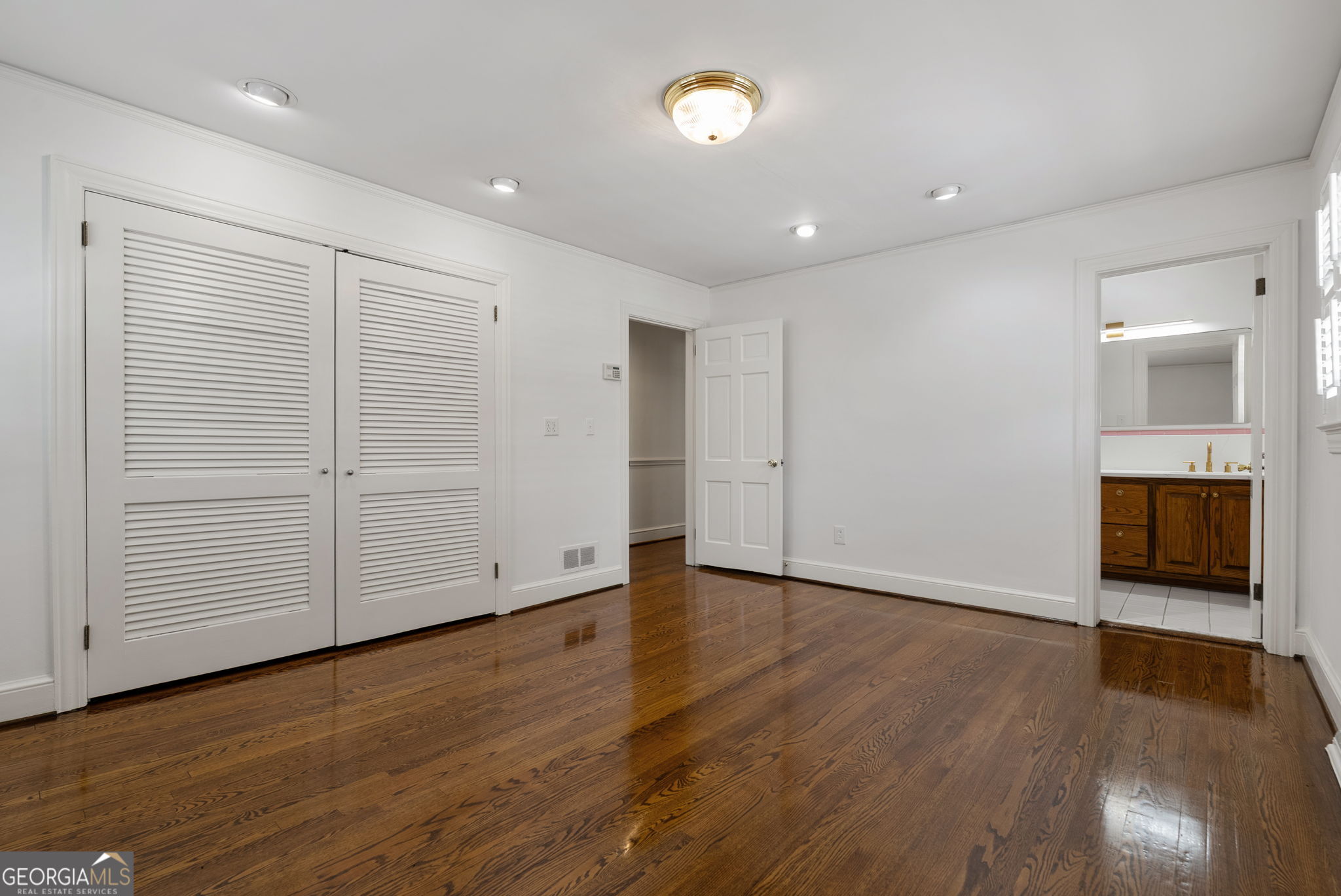 Empty bedroom with polished hardwood floors, louvered closet doors, and a bathroom vanity visible through the doorway.