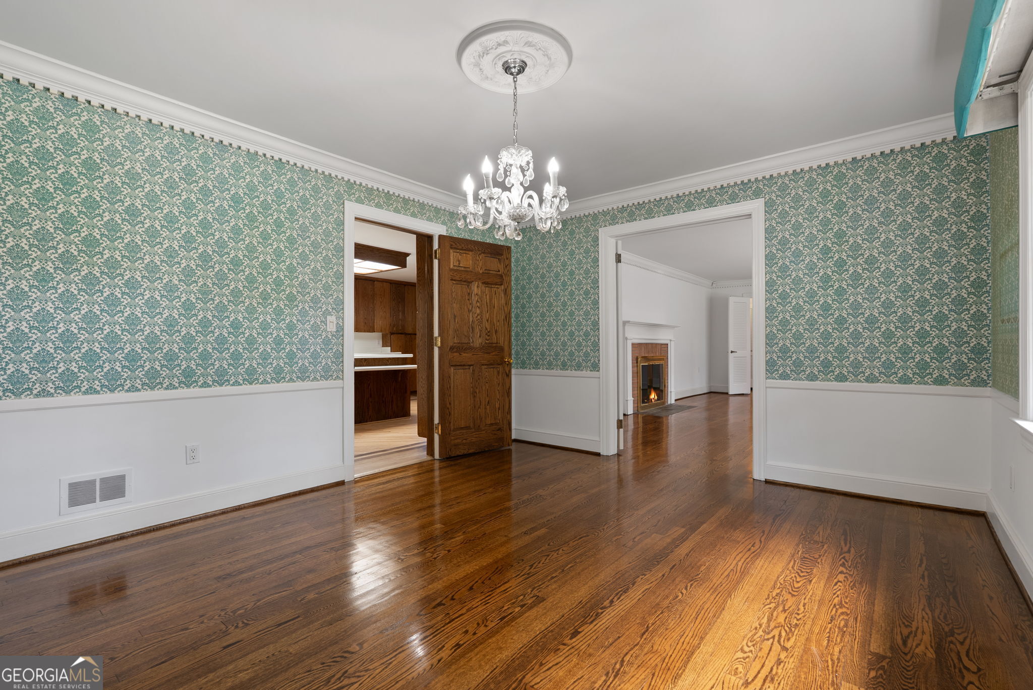 Dining room with teal patterned wallpaper, crystal chandelier, and polished hardwood floors.