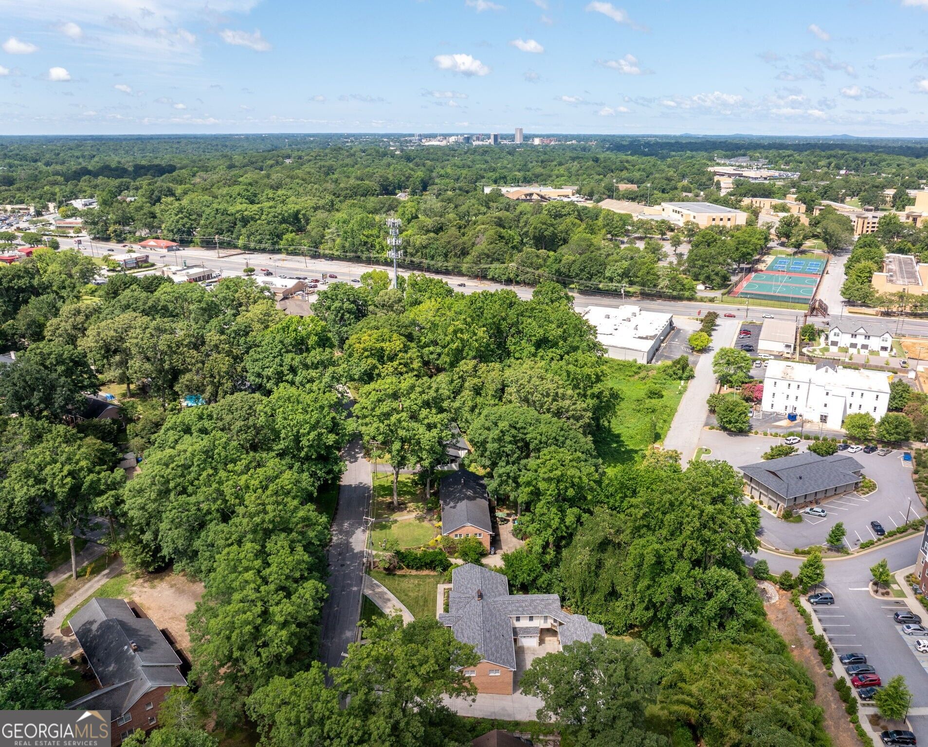 Aerial view of a tree-lined Greenville neighborhood, with a road, tennis courts, and distant skyline.