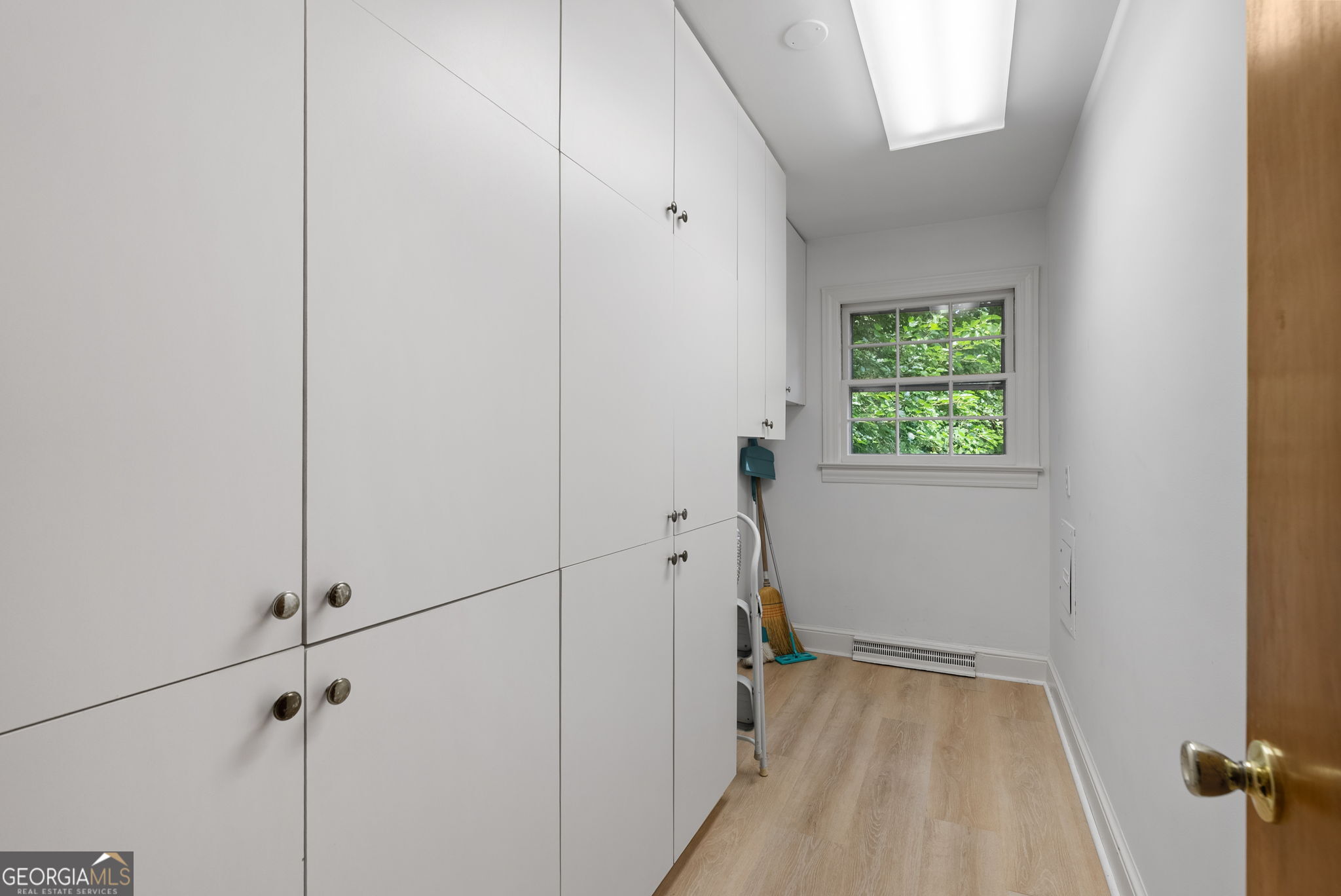 Utility room with white storage cabinets, light wood flooring, and a window overlooking greenery.