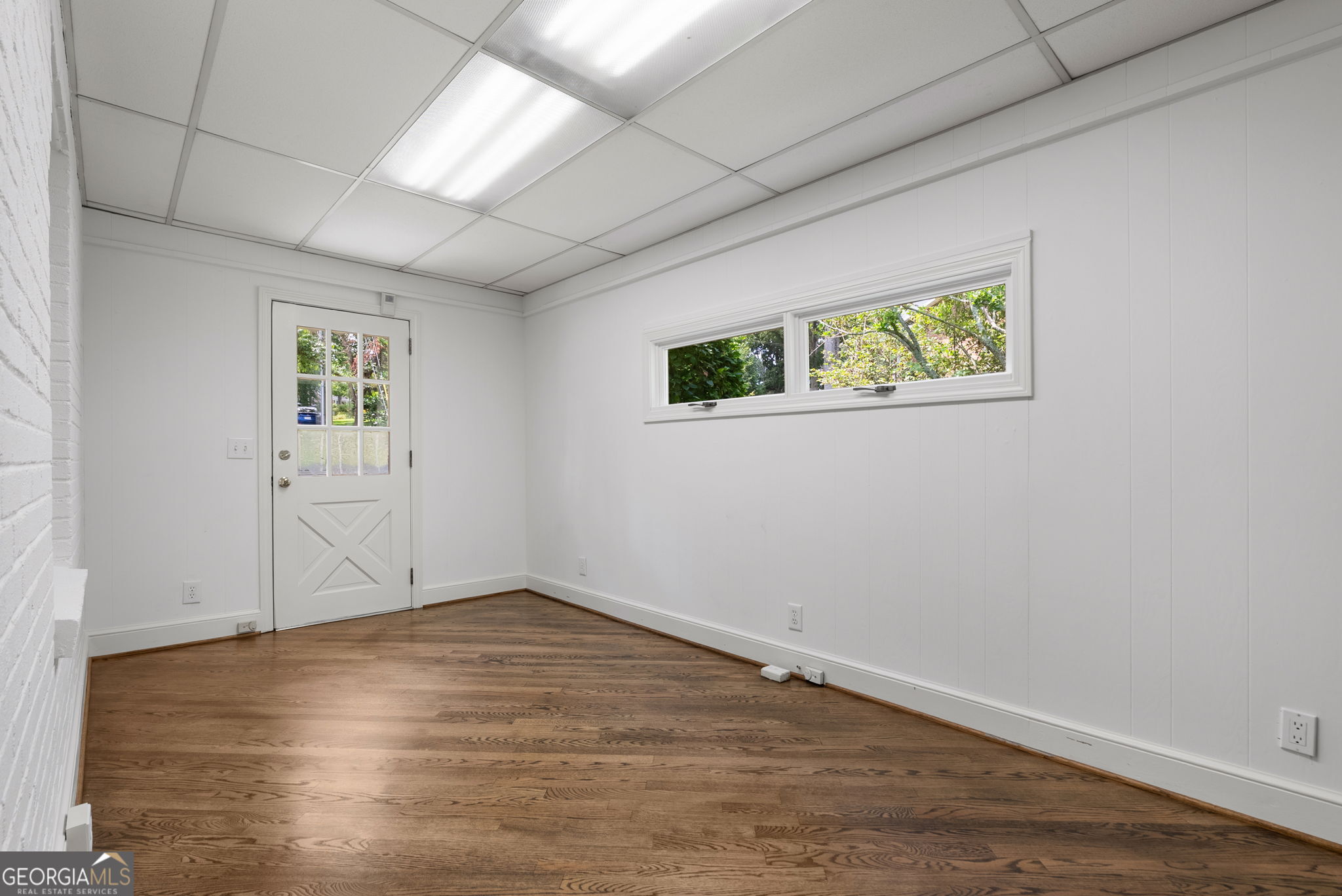 Empty room with hardwood floors, a white brick fireplace, and high windows beside a glass-paneled door.