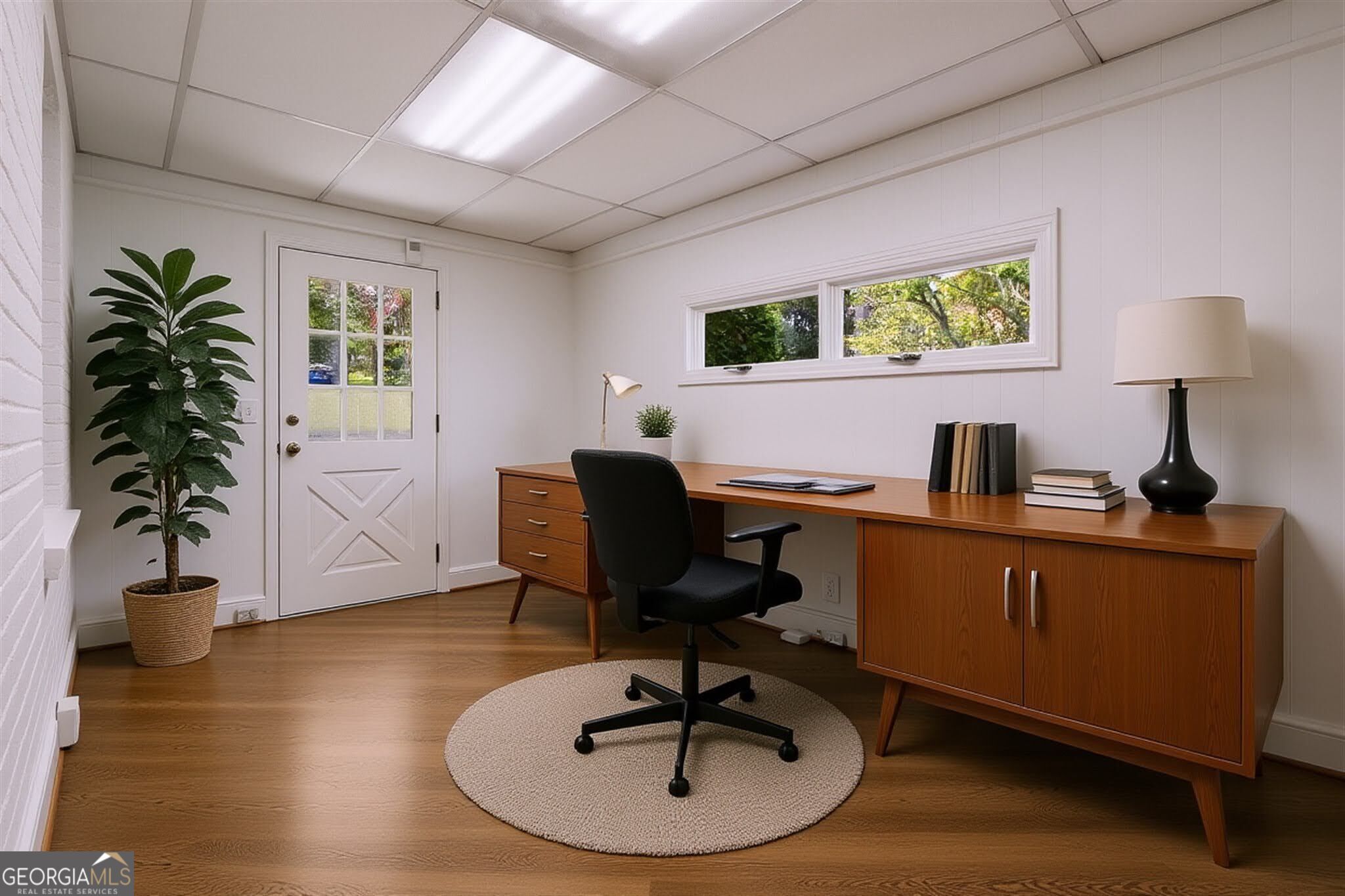 Furnished home office with a long wooden desk, black swivel chair, and narrow windows overlooking trees.