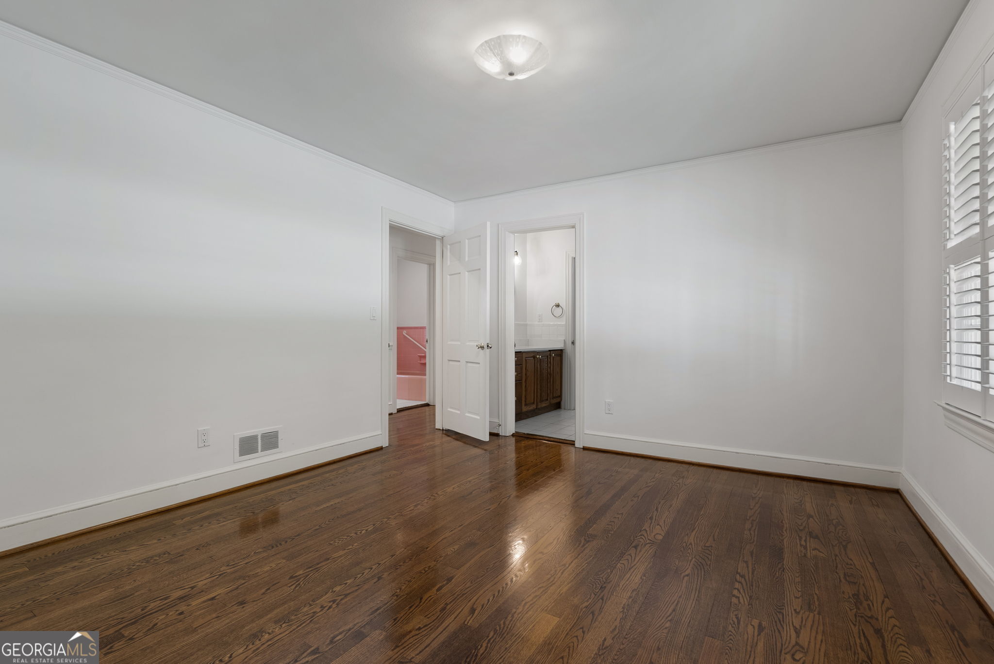 Empty bedroom with dark hardwood floors, plantation shutters, and open doorways to adjoining rooms.