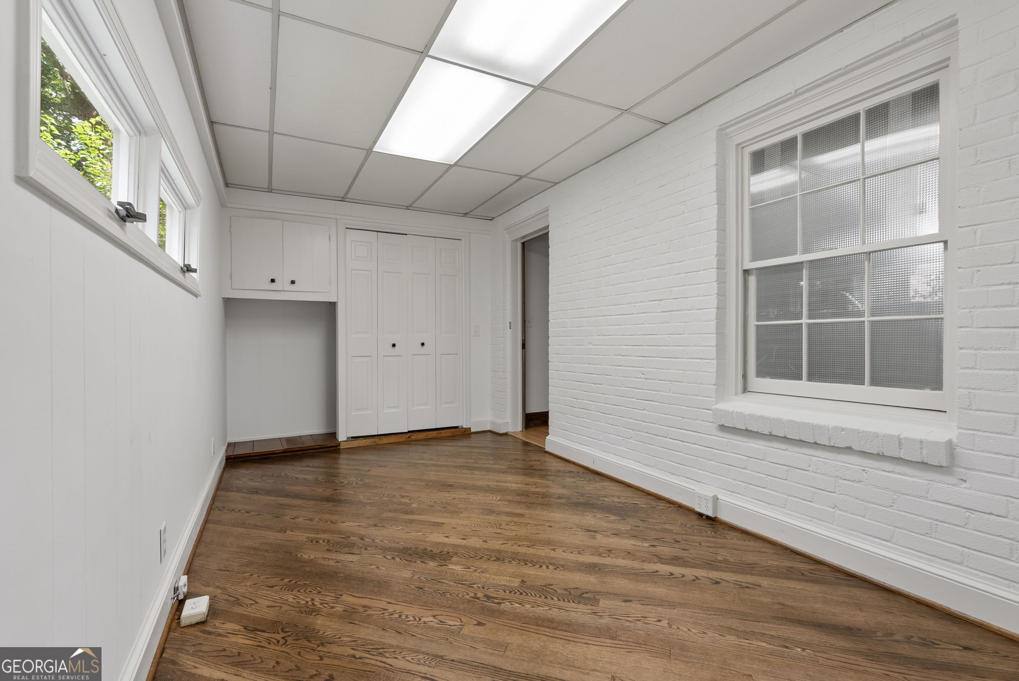 Empty room with hardwood floors, white brick walls, high windows, and built-in cabinets.