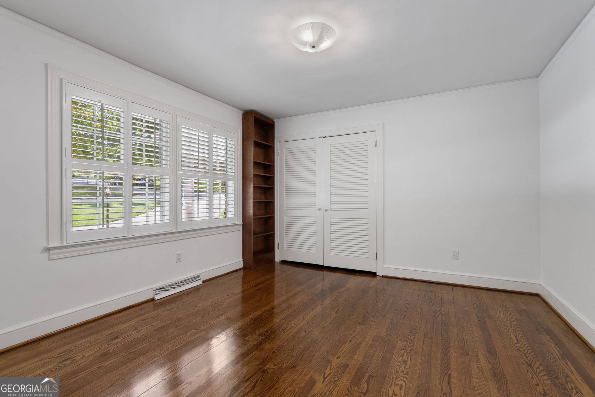 Empty room with hardwood floors, shuttered windows, built-in bookshelf, and louvered closet doors.