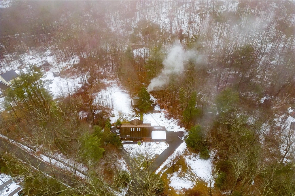 Aerial view of a house in a snowy wooded lot, with smoke rising from its chimney.