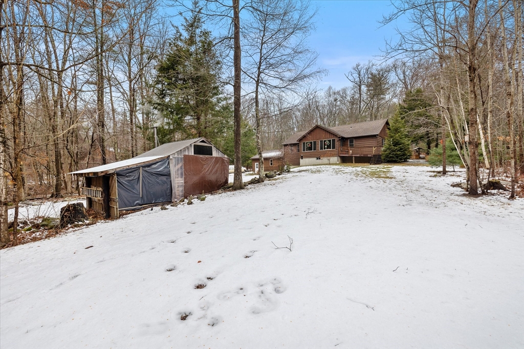 Brown wood-sided house and detached shed on a snow-covered wooded lot.