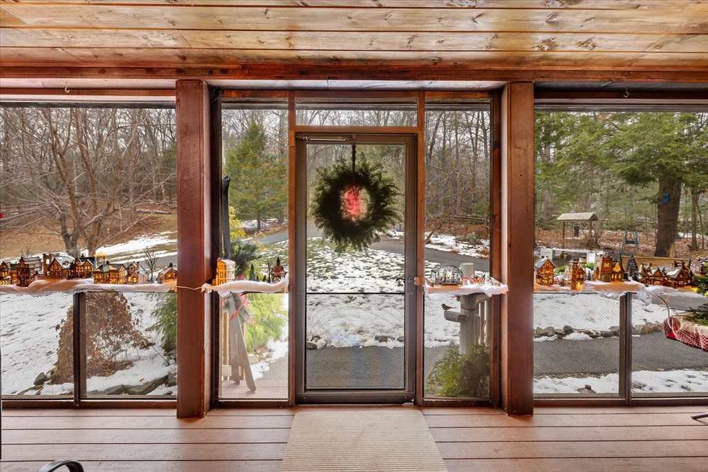 Screened porch with a wreath on the door and miniature village decorations overlooking a snowy wooded yard.
