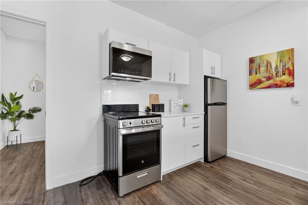 Compact kitchen with white cabinets, stainless-steel appliances, wood-look flooring, and a colorful cityscape painting.