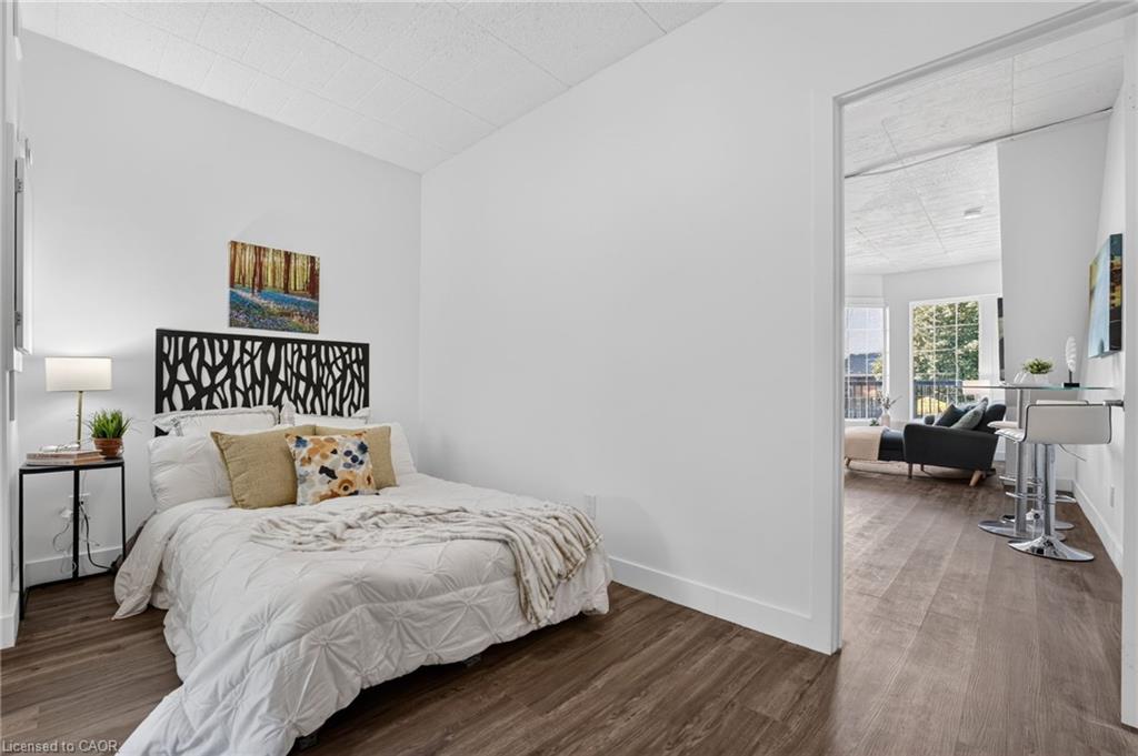Staged bedroom with a white bed and black cutout headboard, opening onto a bright living area.