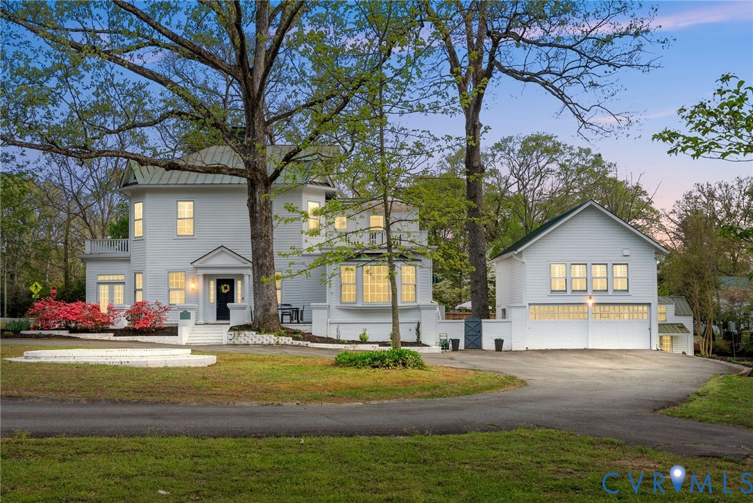 White two-story house and garage beneath mature trees at dusk, with warmly lit windows.