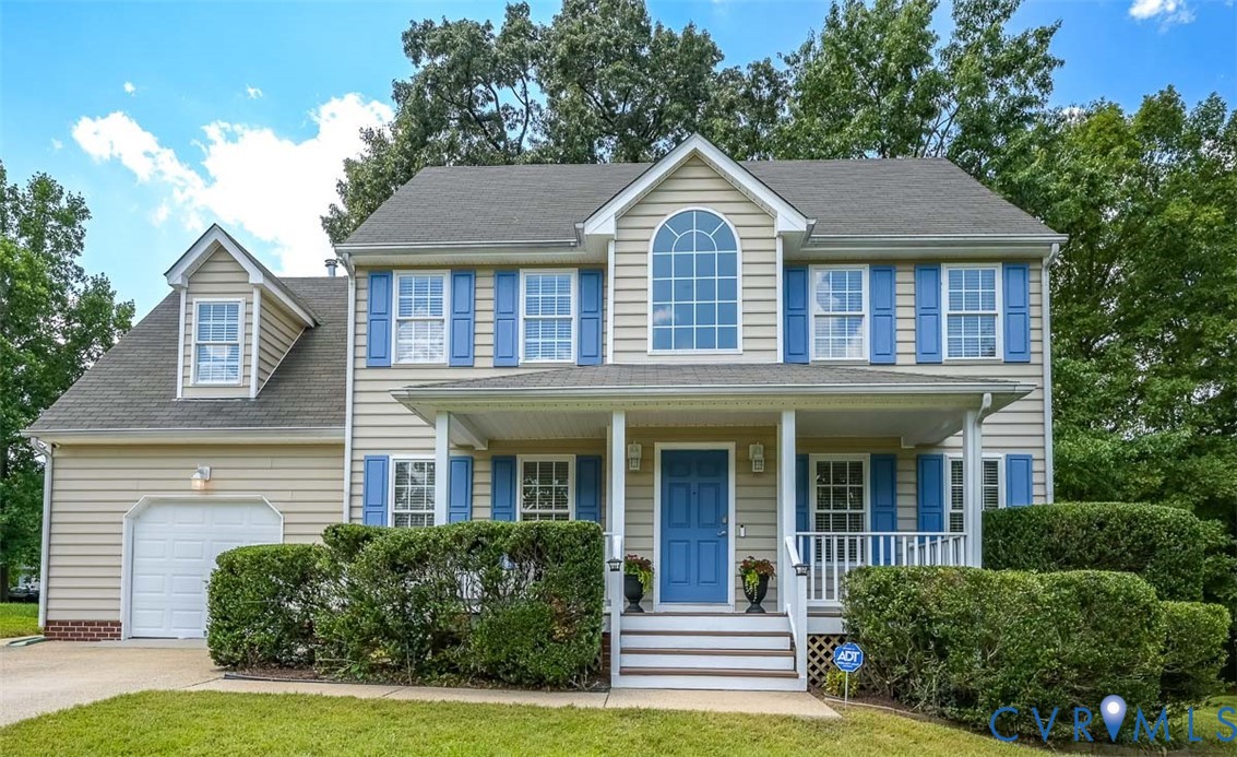 Two-story beige house with blue shutters, a covered porch, attached garage, and neatly trimmed hedges.