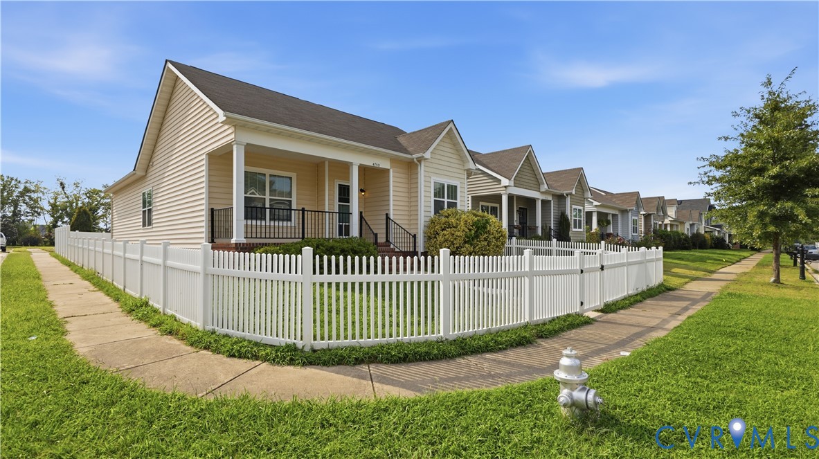 Beige cottage with a covered porch behind a white picket fence, with similar homes along the sidewalk.