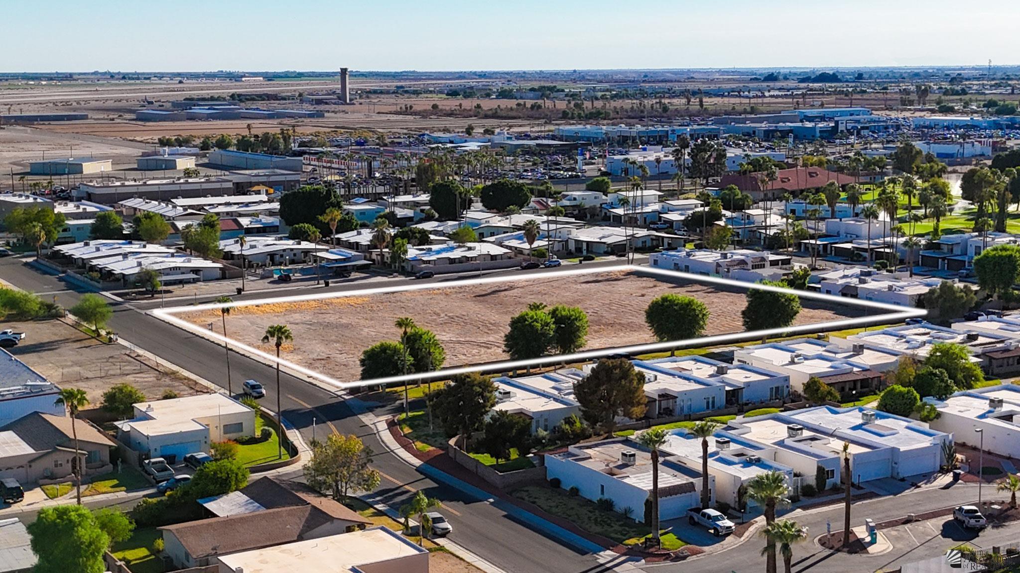 Aerial view of a vacant lot outlined in a residential neighborhood, with commercial buildings in the distance.