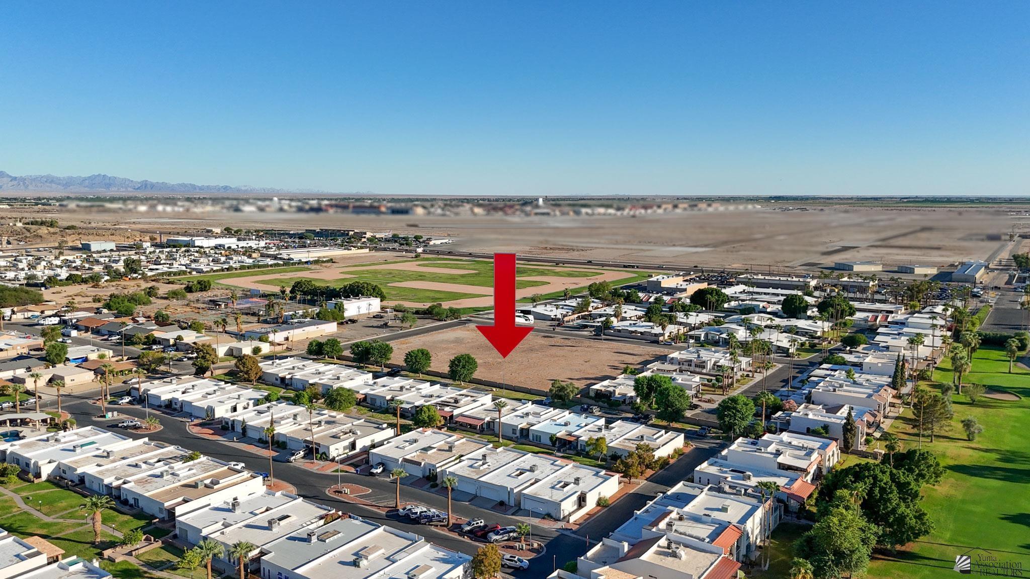 Aerial view of a vacant residential lot marked by a red arrow, near a baseball field and surrounding homes.