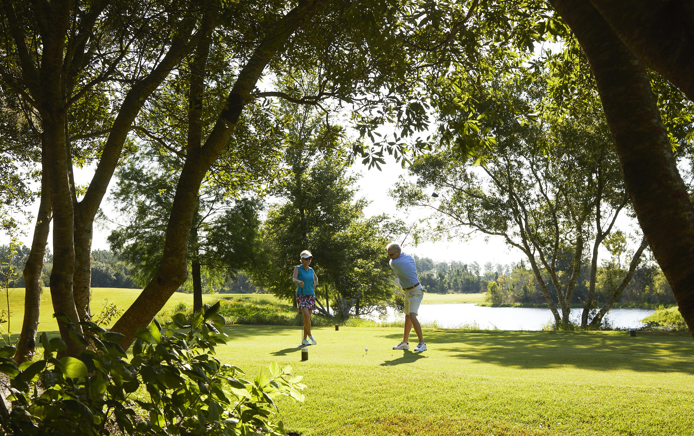 Two golfers play beside a pond on a tree-shaded golf course.