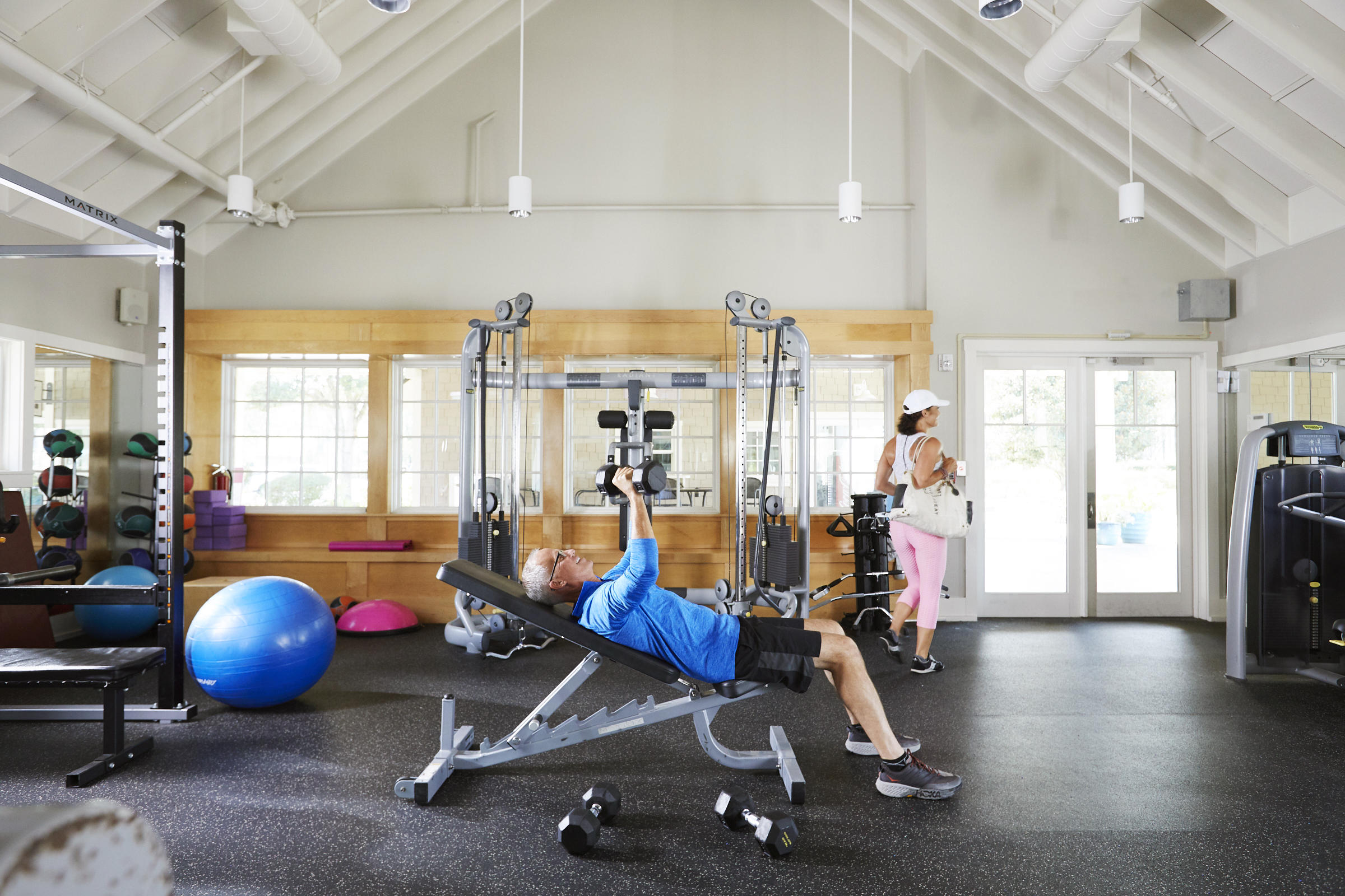 Fitness room with weight machines and large windows; a man lifts dumbbells while a woman stands nearby.