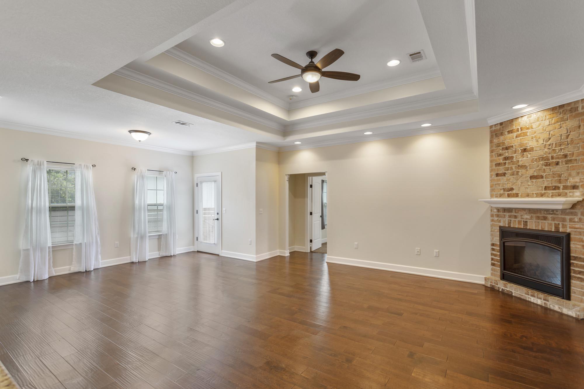 Vacant living room with hardwood floors, a brick fireplace, and a tray ceiling with ceiling fan.