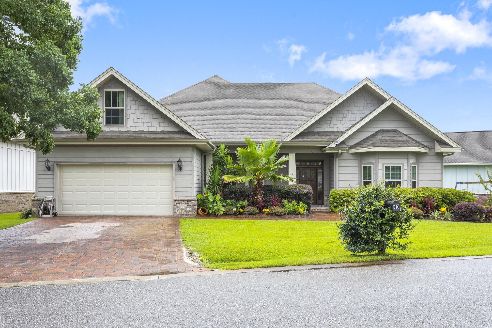 Gray gabled home with a two-car garage, tropical landscaping, and brick driveway.