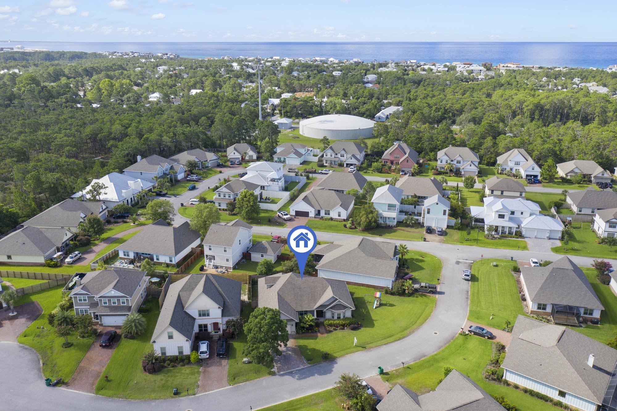 Aerial view of a coastal neighborhood, with the marked home, wooded land, and ocean beyond.
