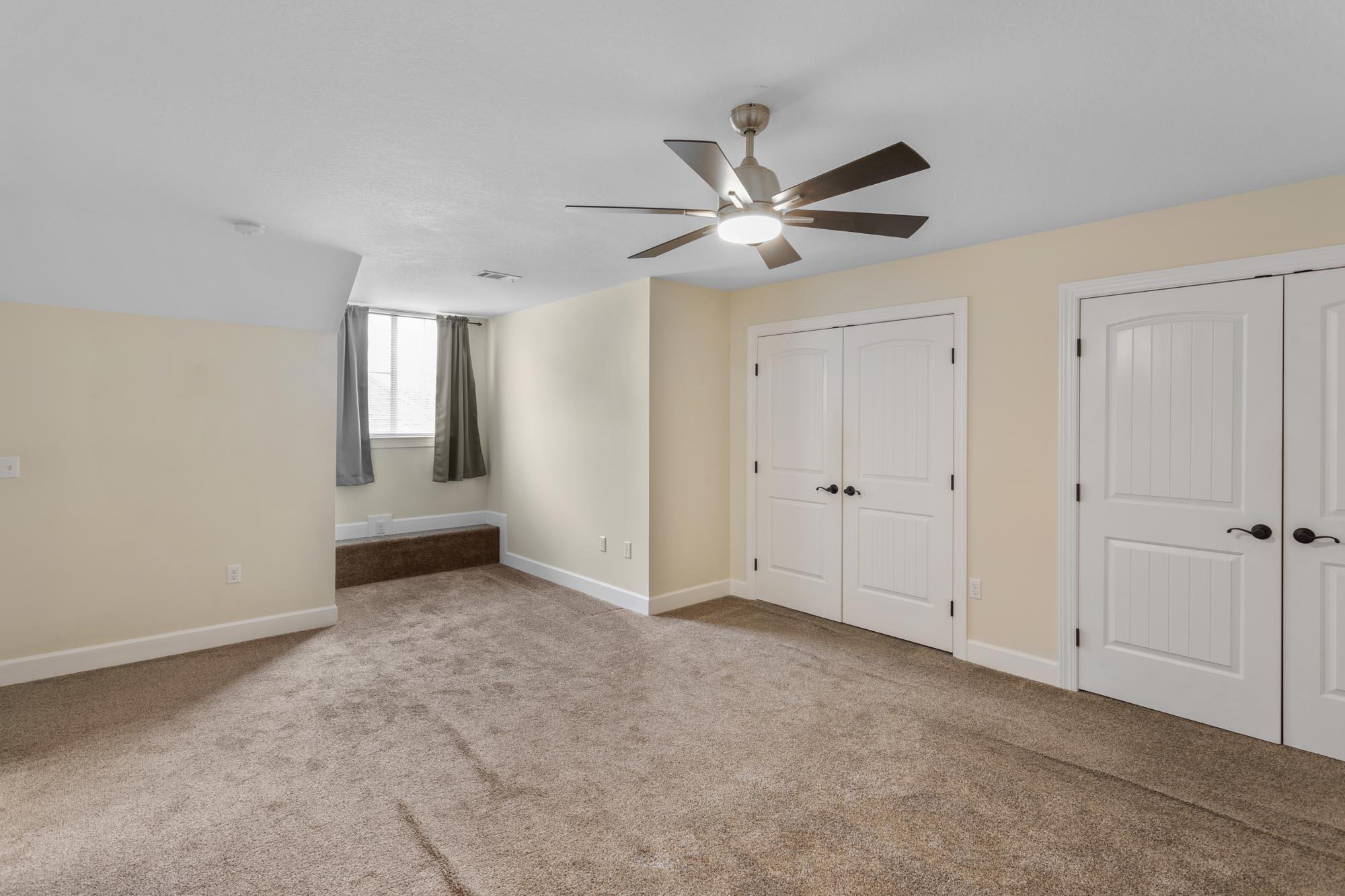 Empty bedroom with beige carpet, a ceiling fan, white closet doors, and a curtained window alcove.