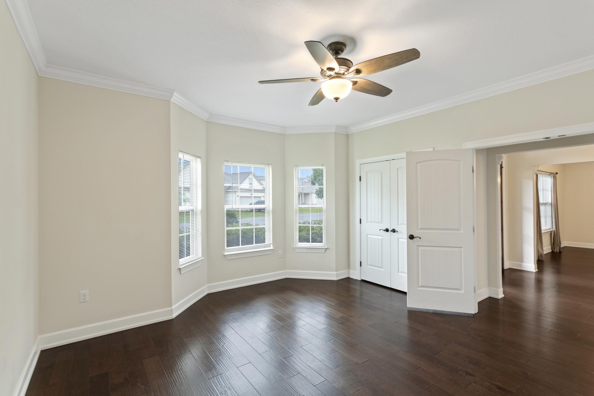Empty room with dark wood floors, three windows, white doors, and a ceiling fan.