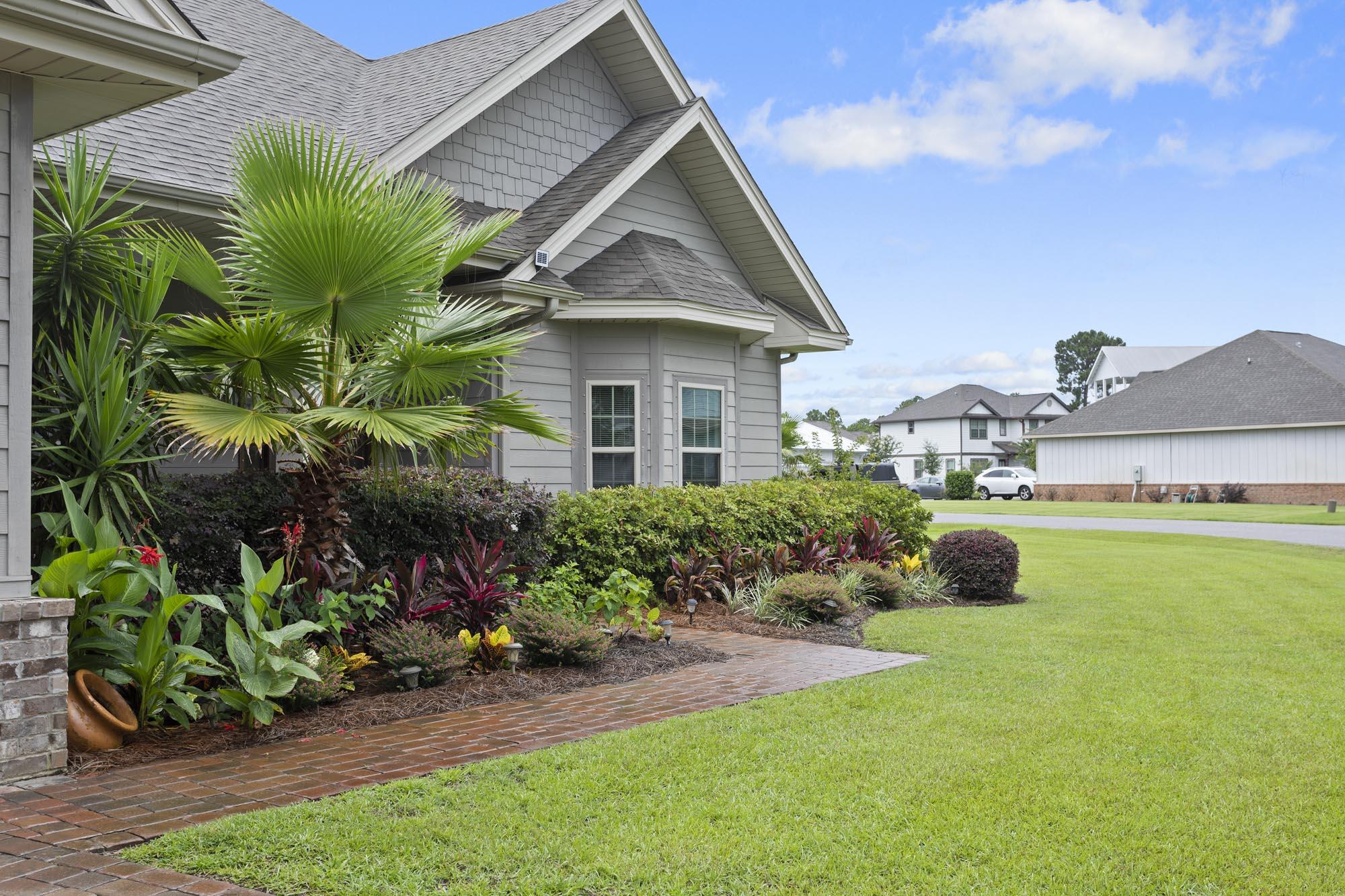 Gray home with tropical landscaping and a brick walkway beside a broad green lawn.