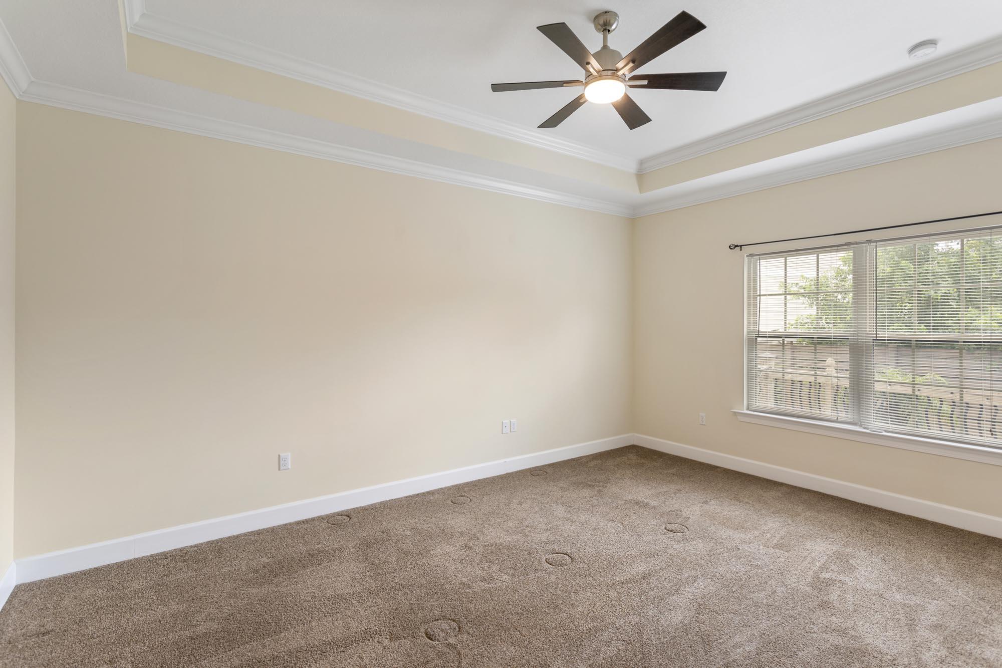 Vacant bedroom with a lit ceiling fan, brown carpet, and a wide window with blinds.