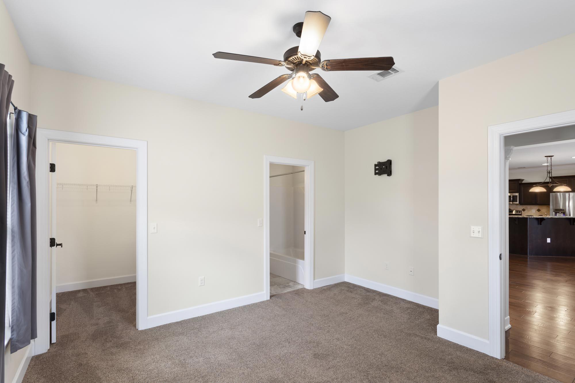 Empty carpeted bedroom with a ceiling fan and doorways to a closet, bathroom, and kitchen.