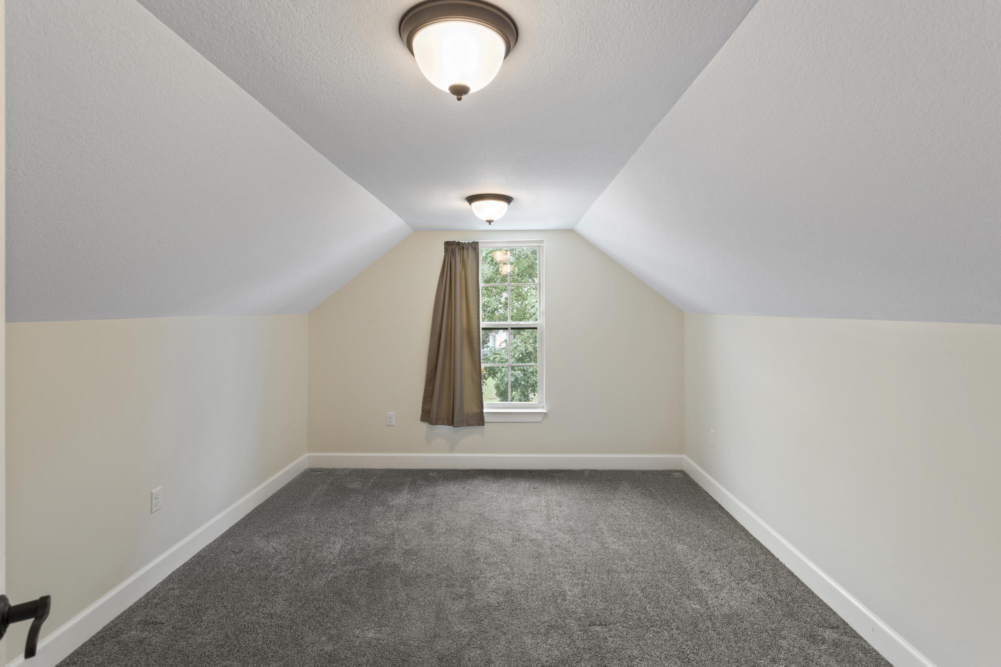 Empty carpeted room with a sloped ceiling and a curtained window.