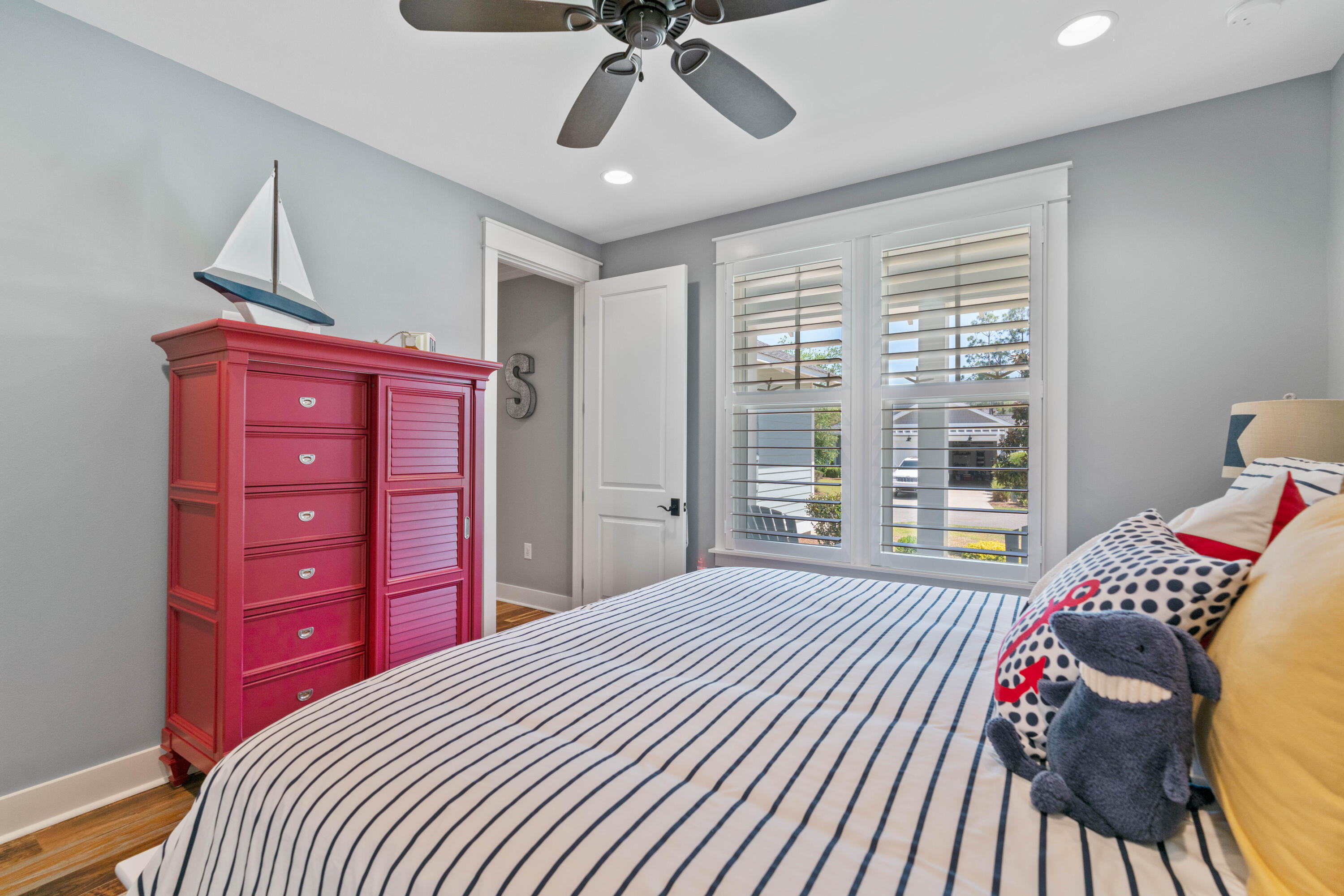 Bedroom with striped bedding, red chest of drawers, sailboat model, and shuttered windows.