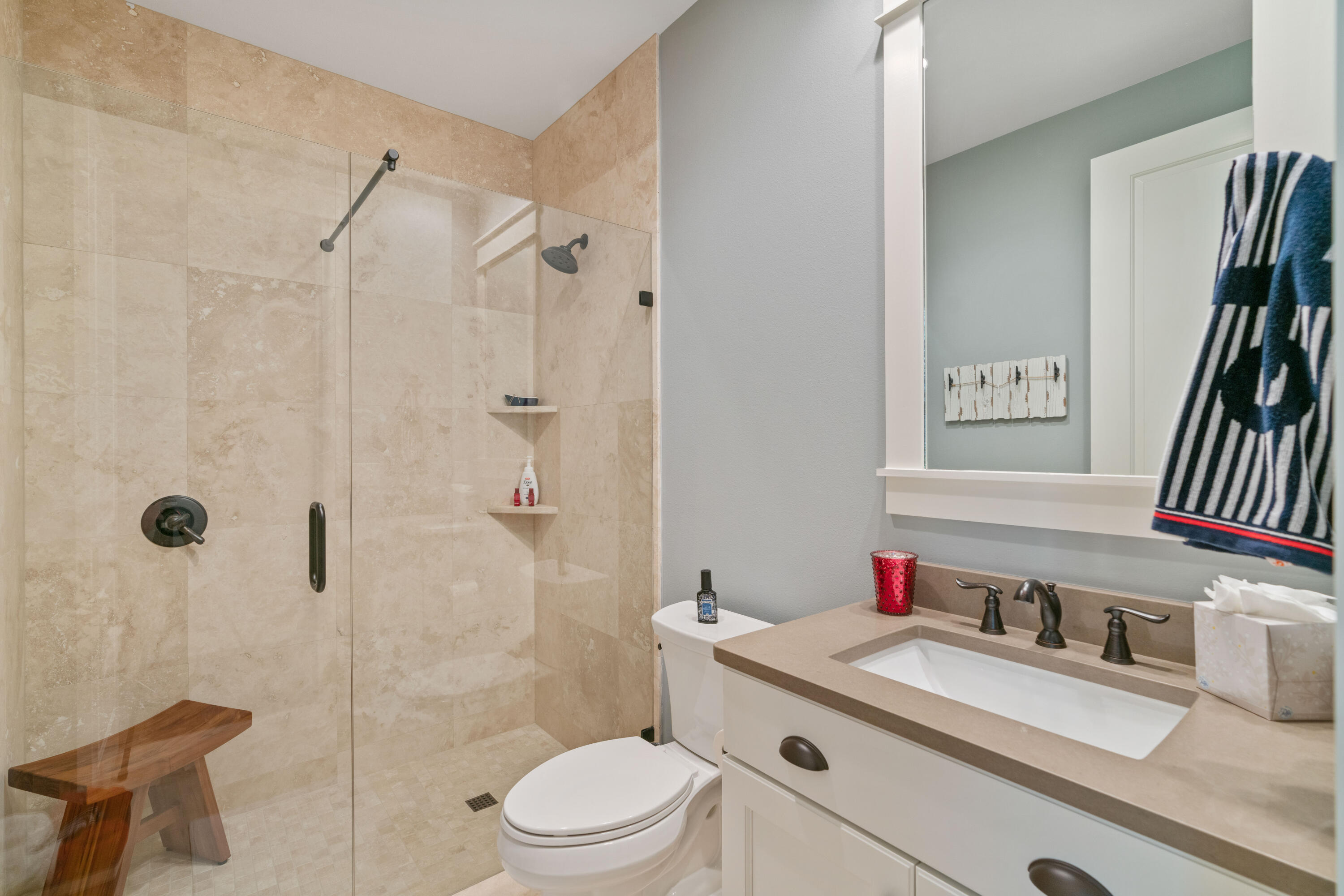 Bathroom with a glass-enclosed beige tile shower, wooden bench, toilet, and white vanity.